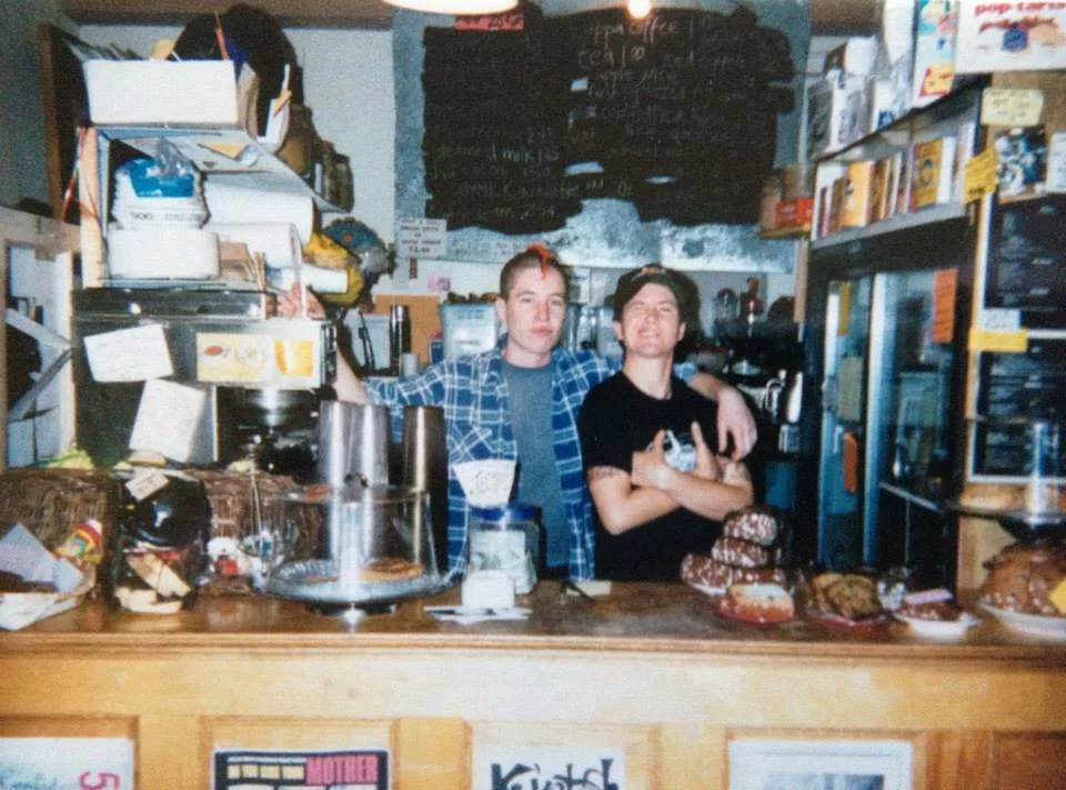 Two masc/butch appearing people with their arms around each other behind a crouded counter and flanked by packed shelves in a cafe. Menu on chalkboard behind them. One is wearing a flanel and has a red tiny mohawk.
