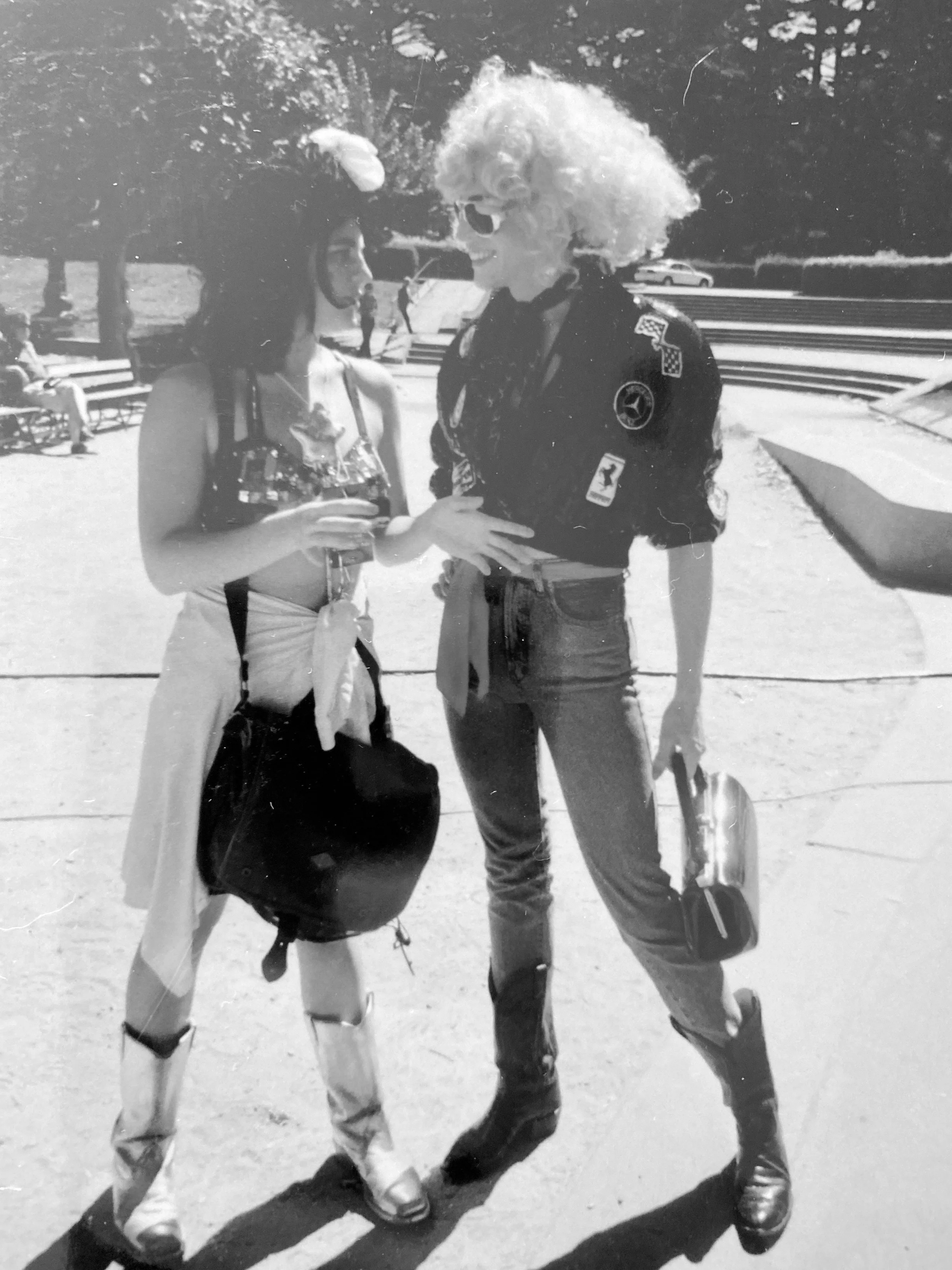 Black and white photo of two fabulously dressed people., wearing boots, crop tops, sun glasses. They are standing close together talking in an outside park