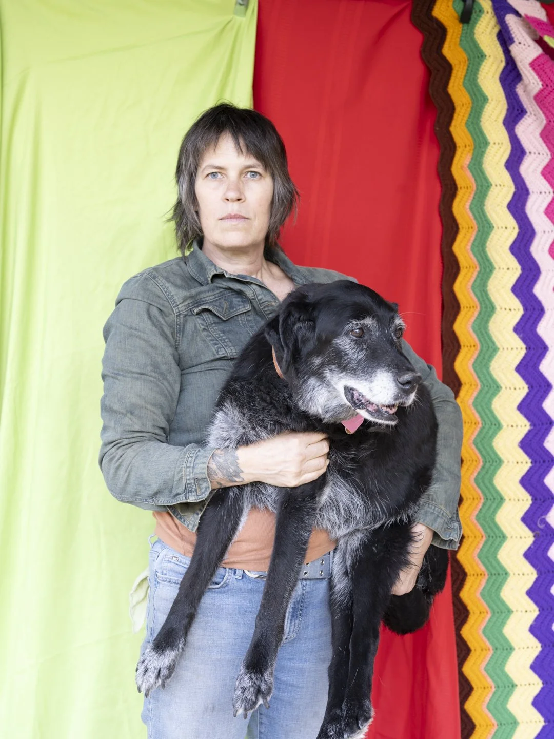 Middle aged, blue eyed, scruffy, bluejean jacket woman, looking serious, holding a 50 pound elderly dog, staring into the distance, with colorful fabric behind her. Mysterious looking