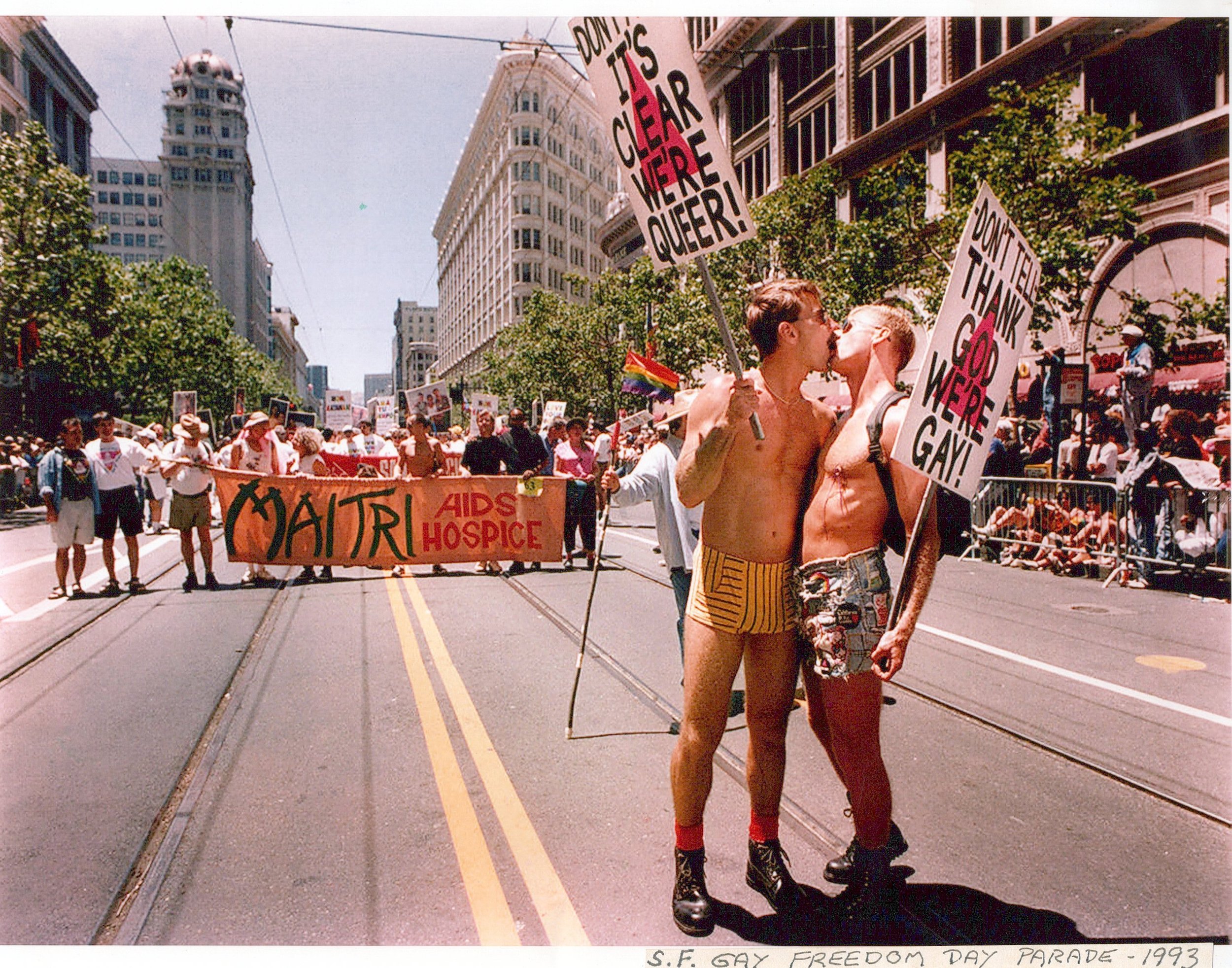 Gay Pride/Freedom parade in SF, background is a line of marchers with Maitri AIDS hospice banner, in the forground is two shirtless men kissing holding large signs that say "It's Clear We're Queer" and "Thank God We're Gay!"