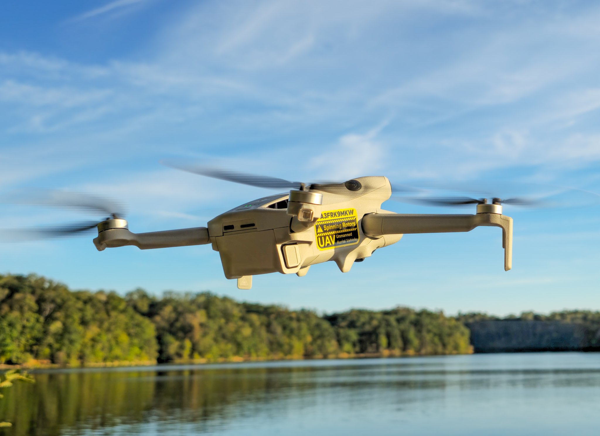 A DJI unmanned aerial vehicle (drone) flying over a body of water with trees and a cliff in the background, under a blue sky with some clouds.