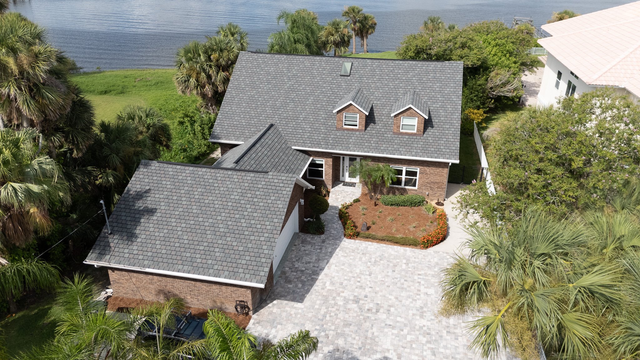 Aerial view of a modern house with gray shingle roof, brick walls, a driveway, surrounded by lush green trees and bushes, with a body of water in the background.
