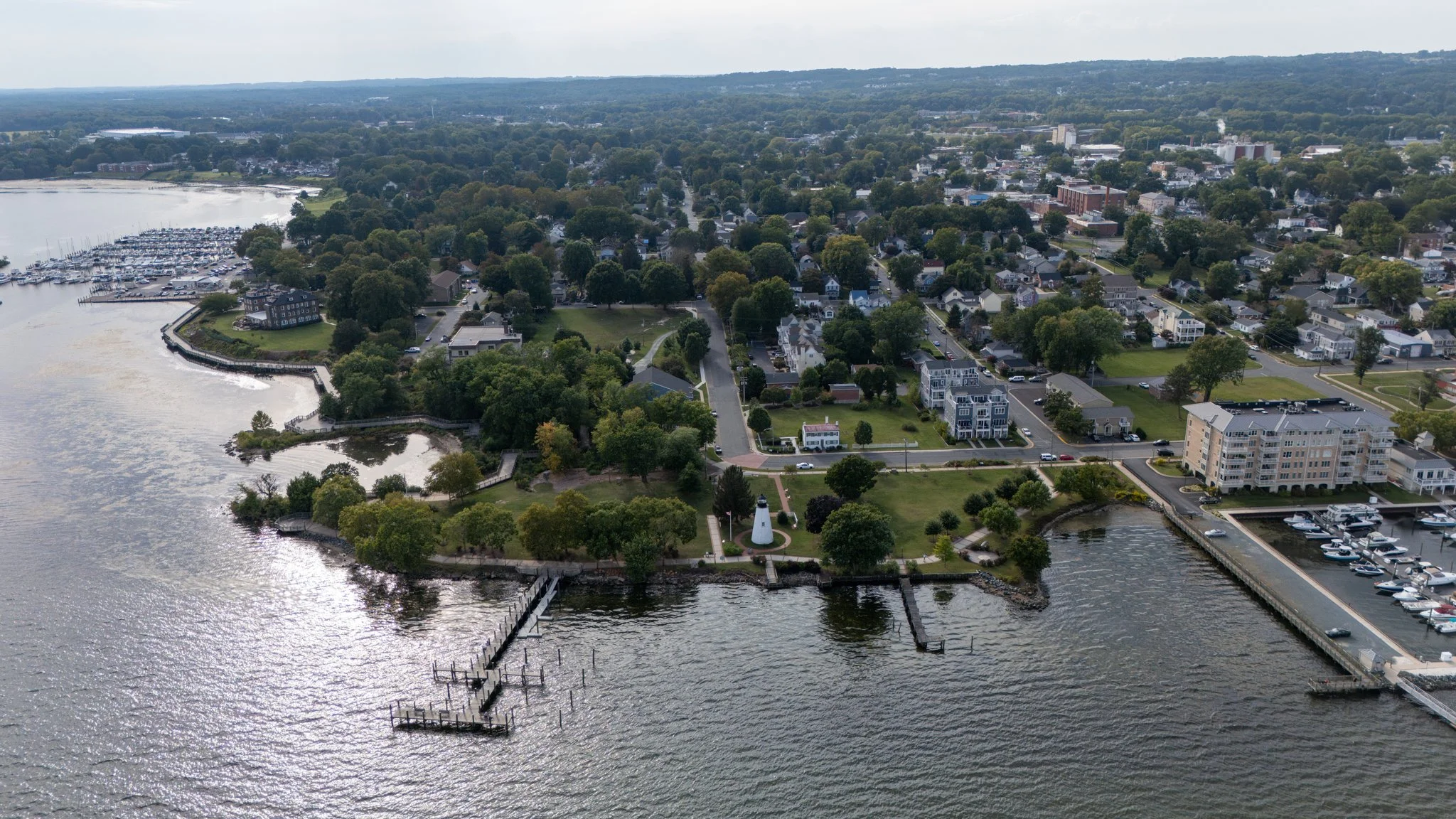 Aerial view of a waterfront town with a marina, lighthouse, residential areas, and green spaces.