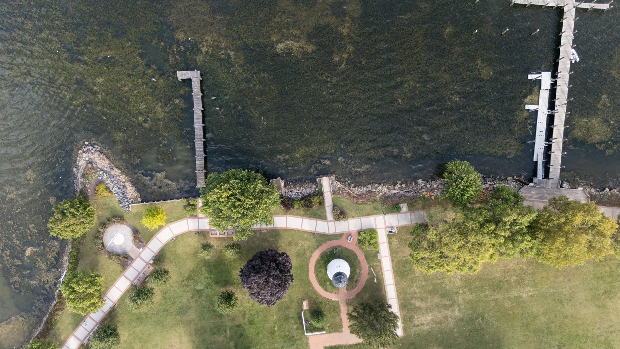 Aerial view of a park with a walking path, trees, grass, and a waterfront with docks and a lighthouse.