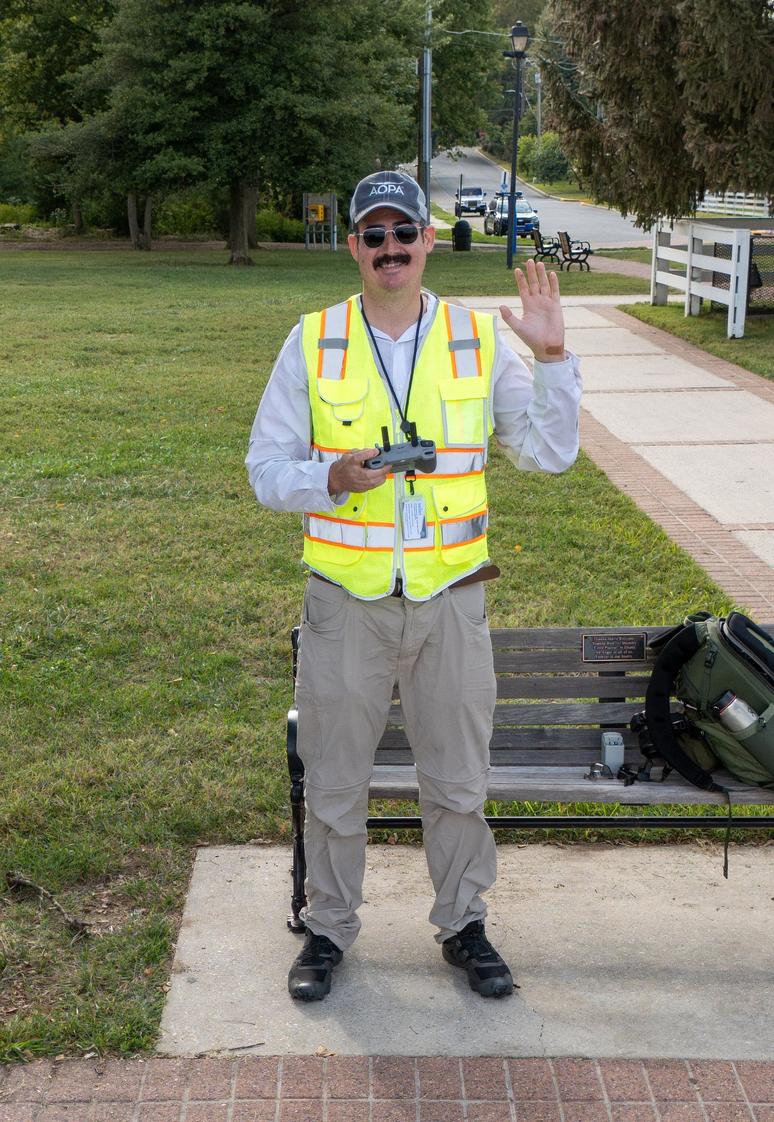 A man wearing a reflective safety vest, sunglasses, and a cap, standing outdoors on a sidewalk, holding a remote control, and waving. There is a park bench with a backpack and some electronic devices beside it. The background shows trees, a street, and parked cars.