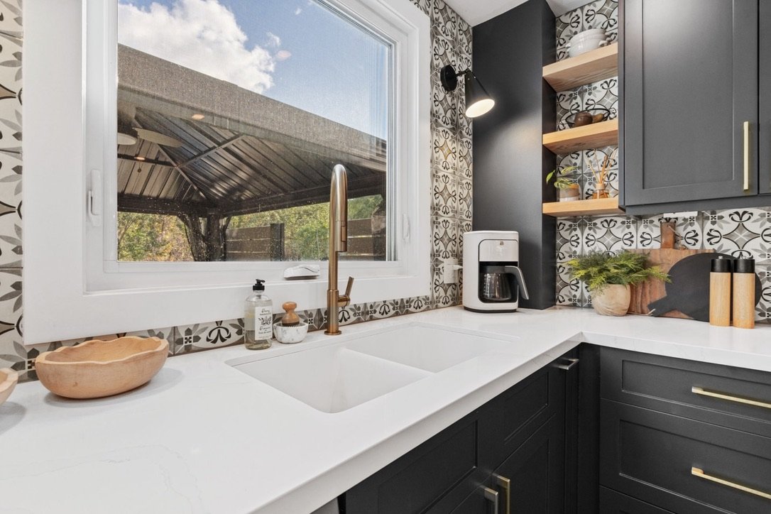 Kitchen with black cabinets, a white countertop, patterned backsplash, and a large window showing outdoor trees and a covered patio.