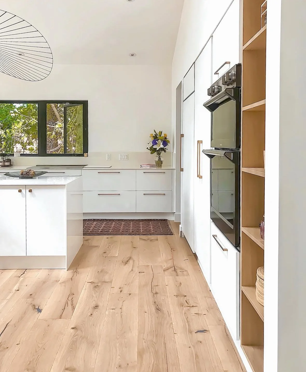 Bright kitchen with white cabinets, wooden shelves, and a black window overlooking trees. A vase with flowers on the countertop. Wooden flooring and a rug near the counter.