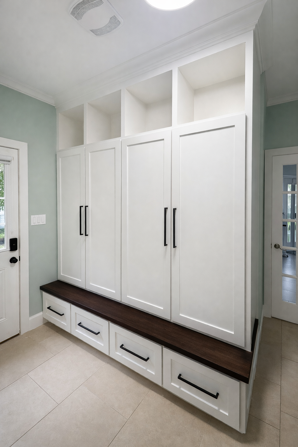 White built-in storage cabinets with black handles, a dark wood bench, and open cubbies on top, in a room with beige tile flooring and green walls. A door with a glass panel is on the right, and a white door with a black handle is on the left.
