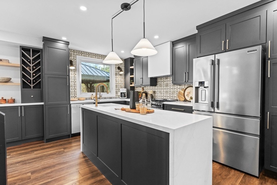Modern kitchen with dark gray cabinets, white countertops, stainless steel refrigerator, patterned backsplash, wooden open shelves, and a kitchen island with two white pendant lights.