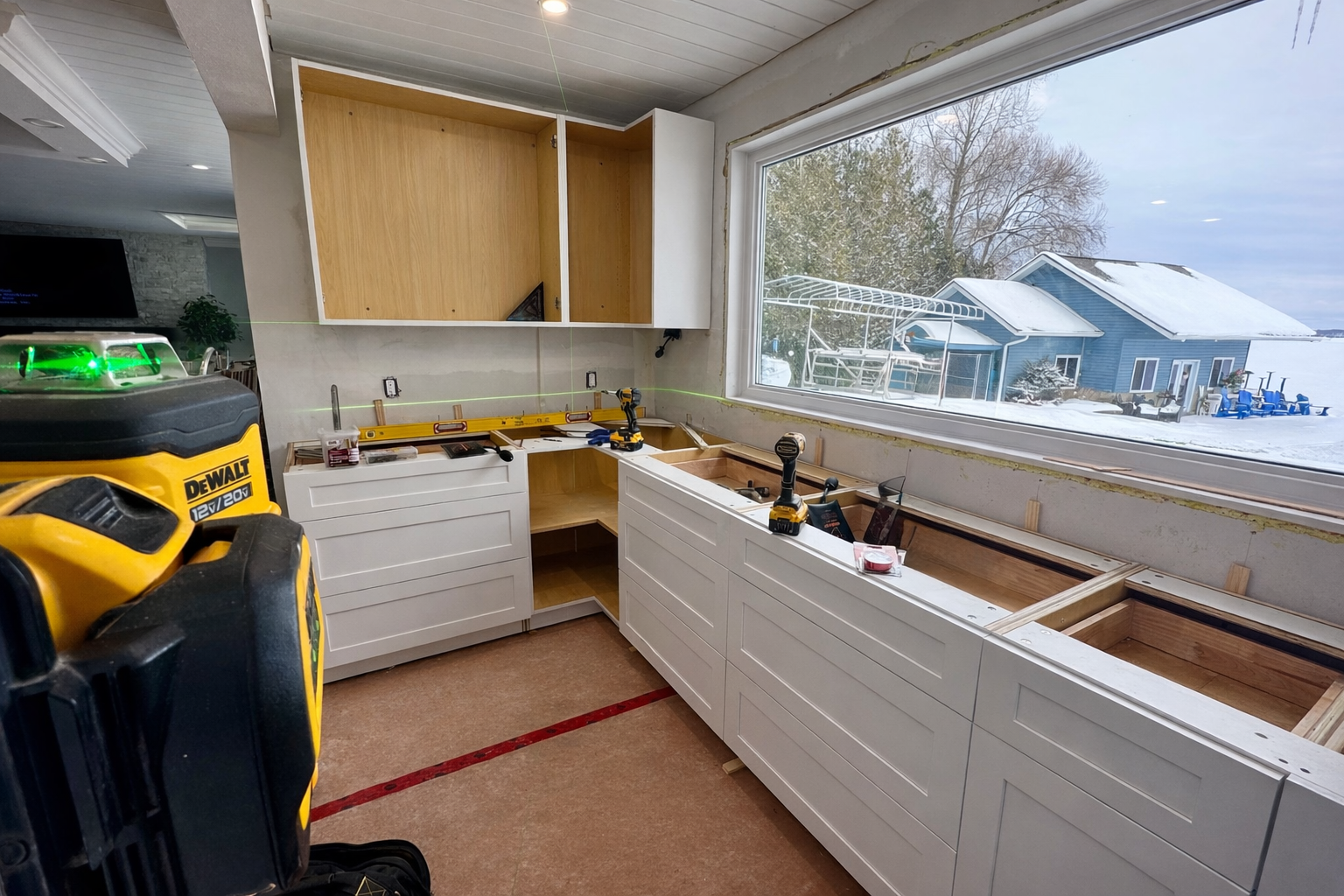Kitchen renovation under construction with white cabinets, construction tools, and a large window showing a snowy outdoor scene with a blue house and outdoor furniture.