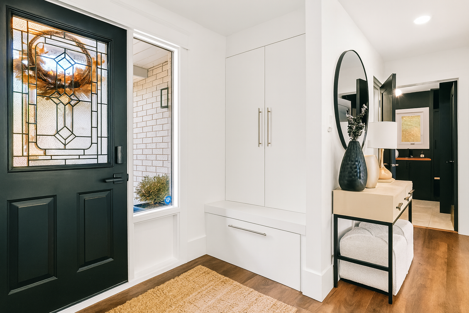 Entryway with black front door featuring stained glass window, white walls, built-in white storage cabinet, and a beige side table with decorative vases and a mirror.
