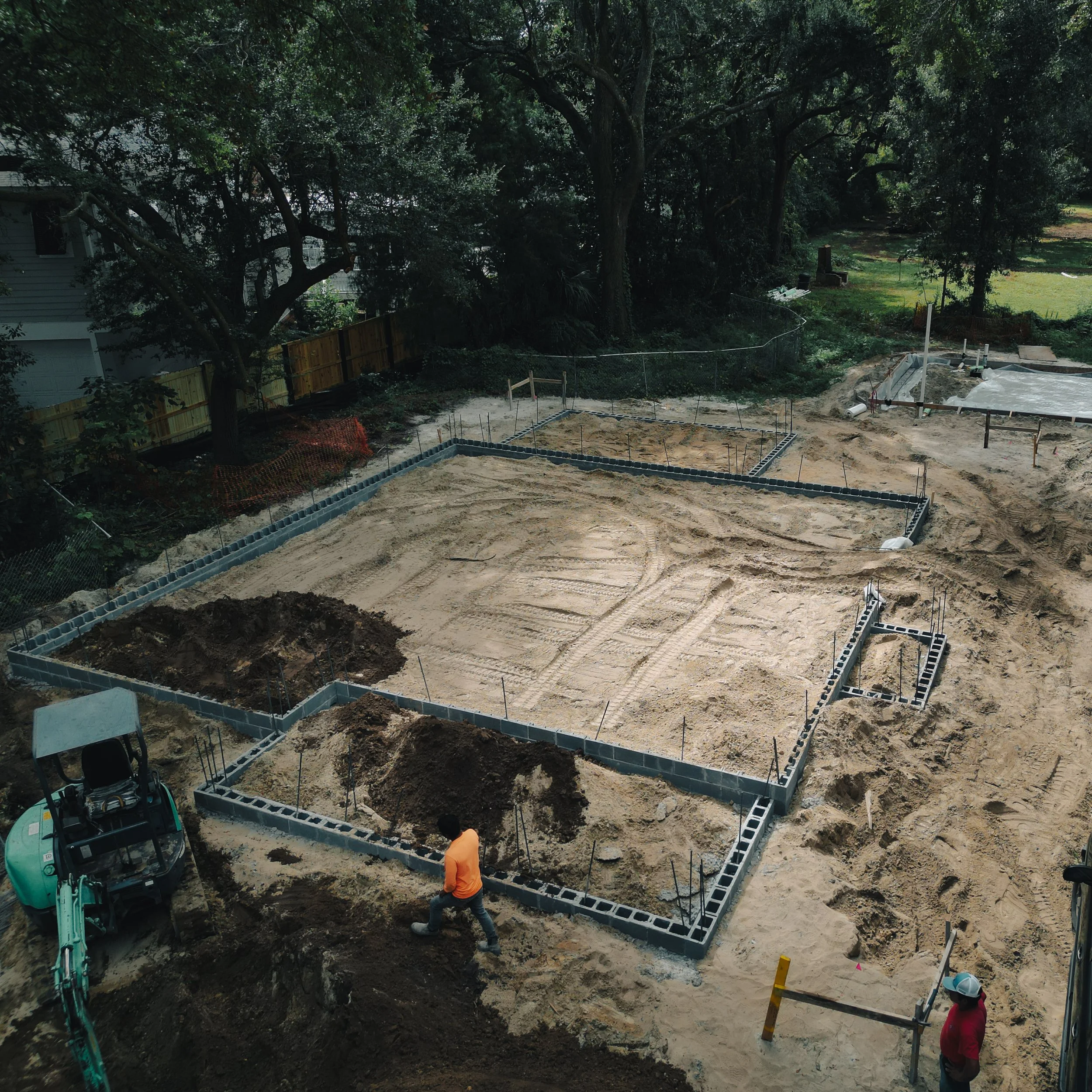 Construction site with foundation being laid, concrete blocks forming the outline of a building, and two workers present.