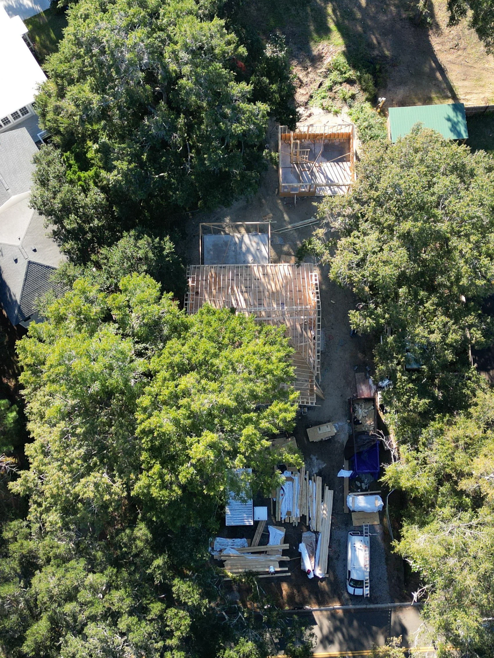 Bird's eye view of a house under construction with wooden framing, surrounded by green trees, construction materials, trucks, and a driveway.