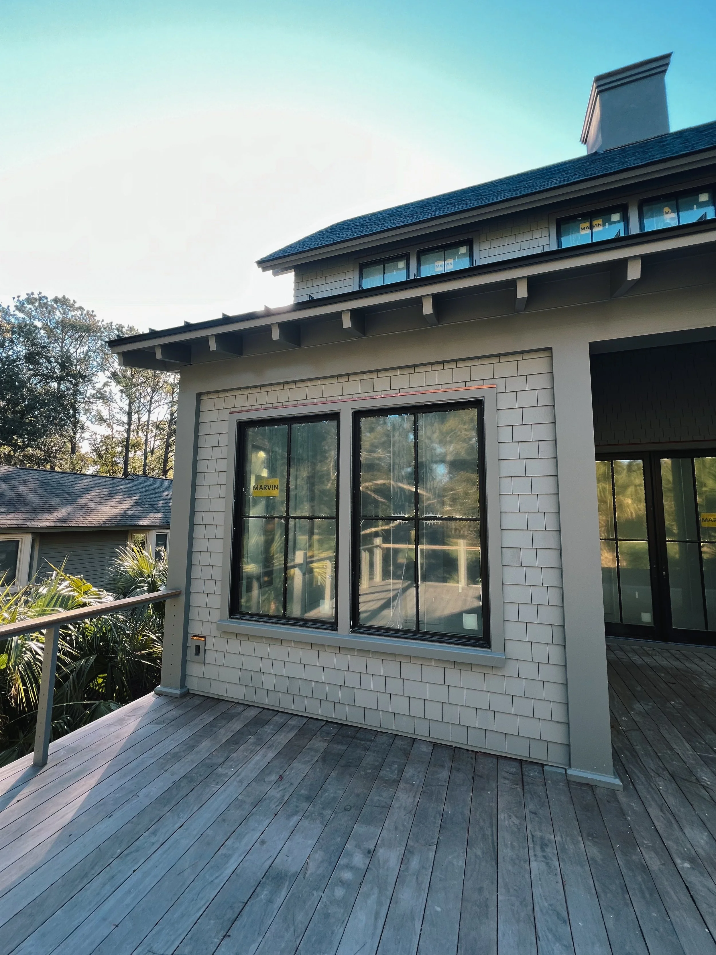 A new two-story house with a wooden deck, large double-pane glass windows, siding made of white shingles, and a roof with exposed rafters. The house appears to be under construction or newly built.