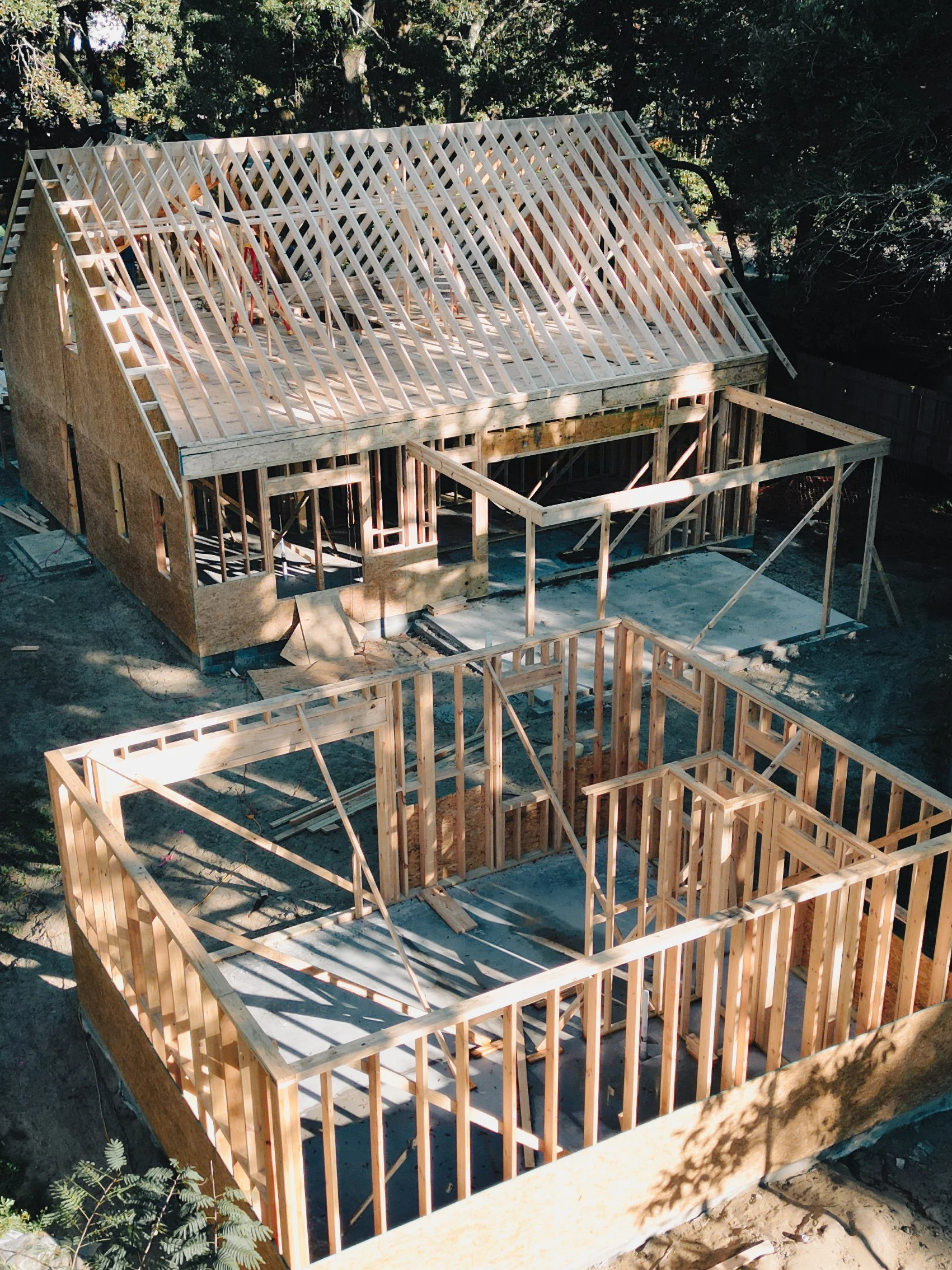 Two wooden house frames under construction, showing the framing of the walls and roof with sunlight filtering through trees.