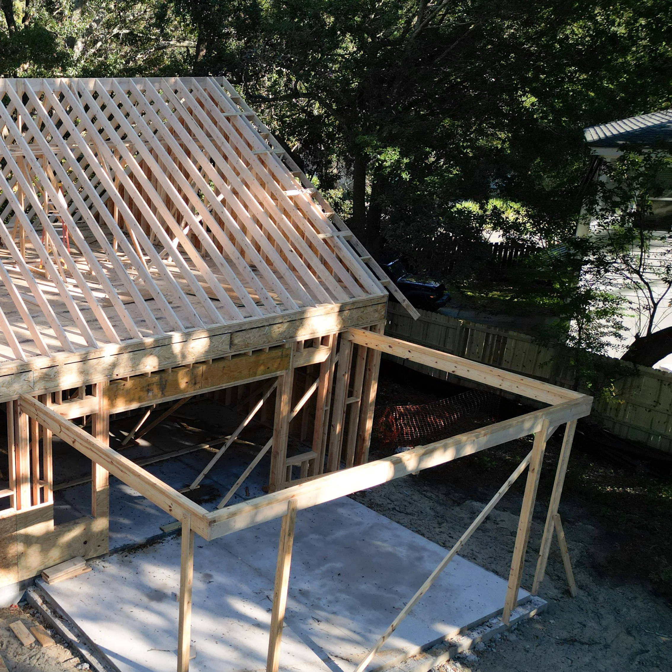 Wooden frame of a house under construction, with roof trusses installed, on a concrete foundation in a backyard with trees and a fence.