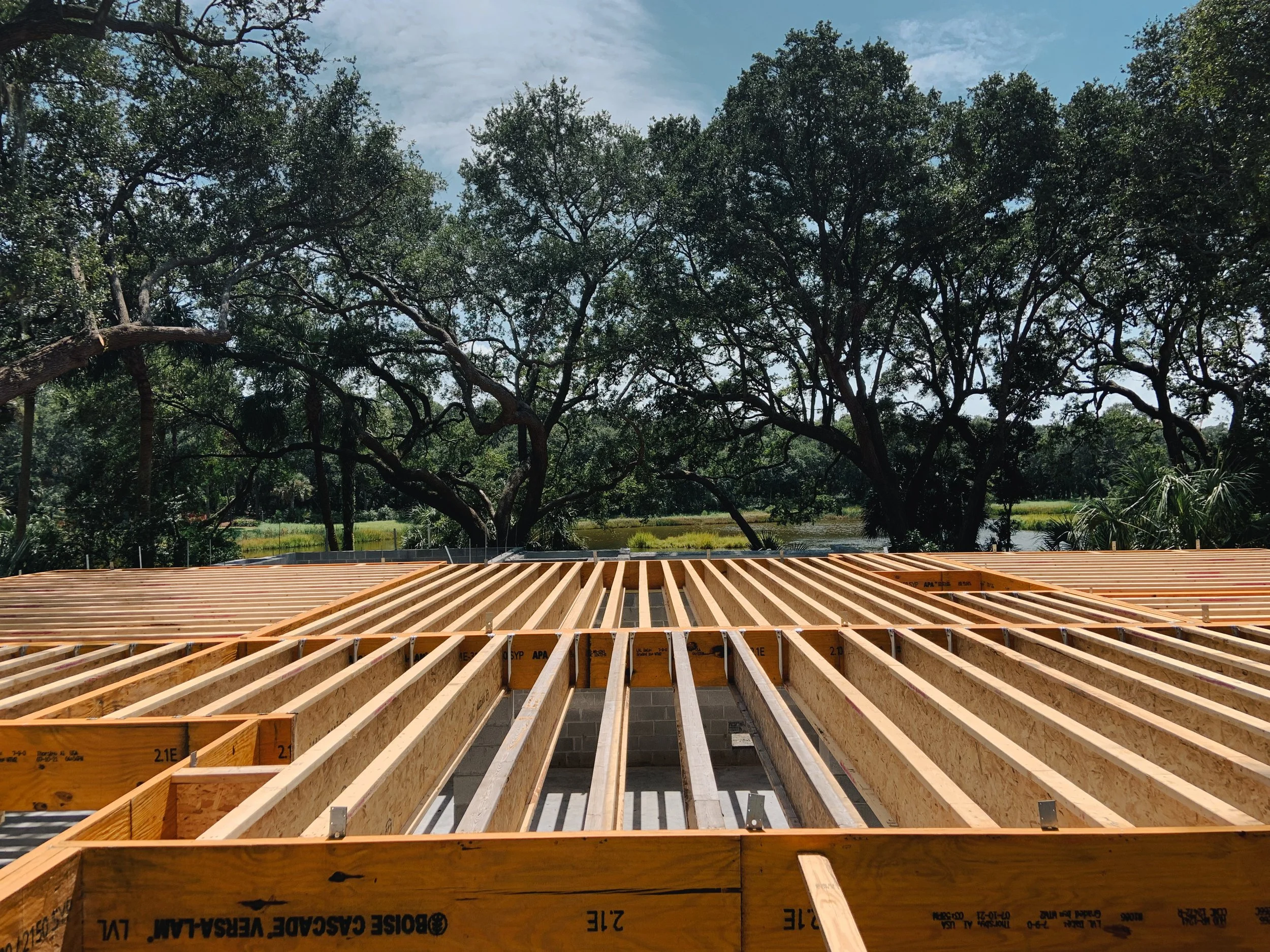 Wooden framing construction on a building in a wooded area with large trees and a blue sky.