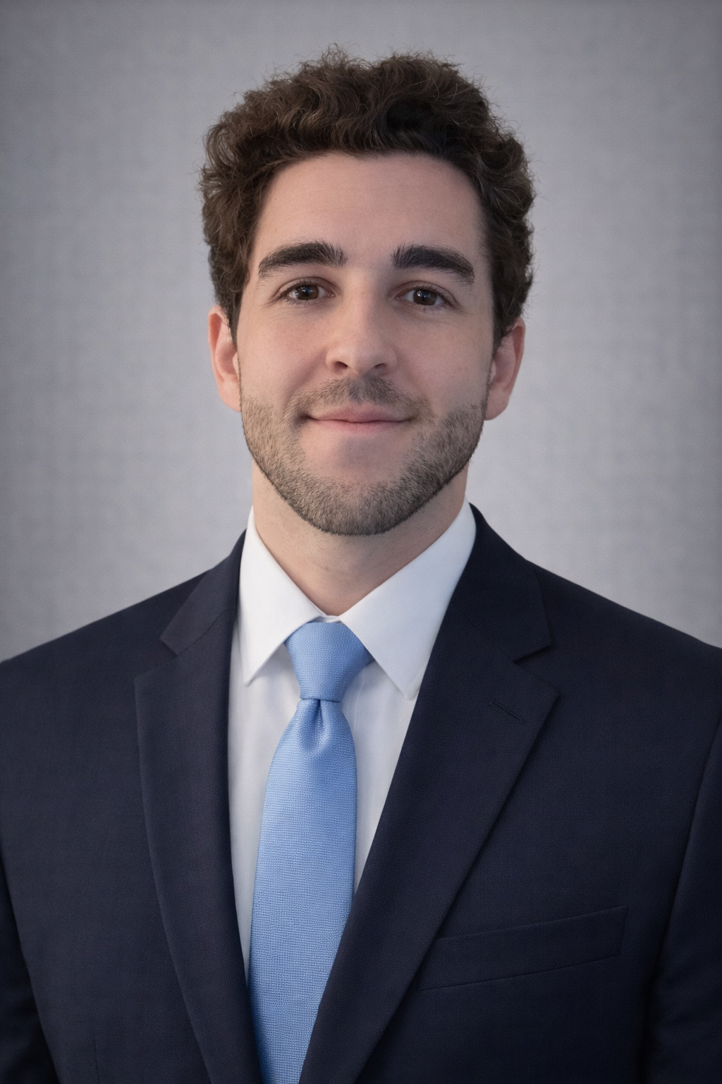 Headshot of a young man in a dark suit, white shirt, and light blue tie, smiling softly against a neutral background.