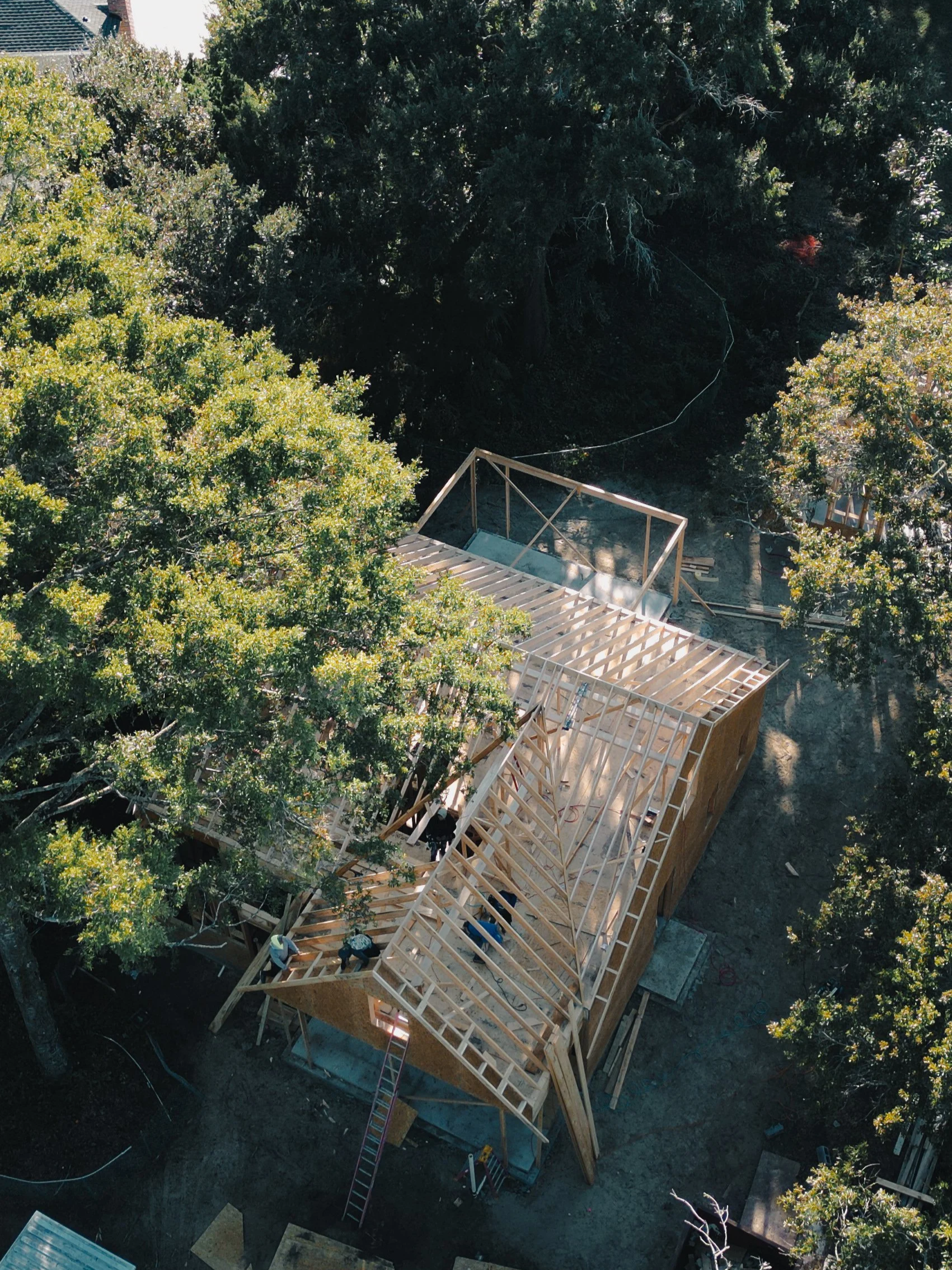 Aerial view of a house under construction surrounded by trees, showing wooden framing and roof structure.