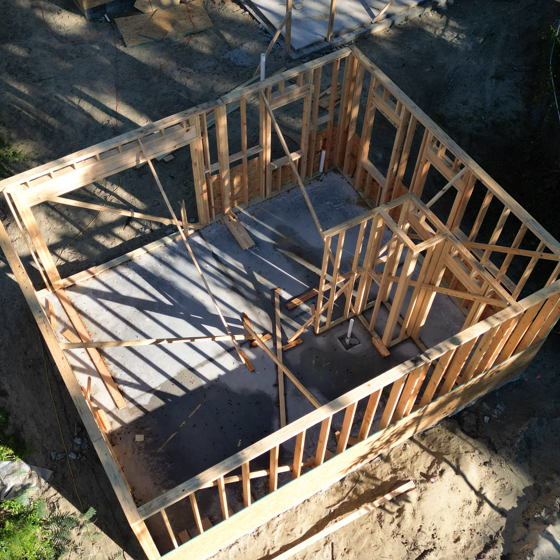 Wooden framework of a house under construction with visible walls and partial roof framing, on a concrete foundation with dirt around.