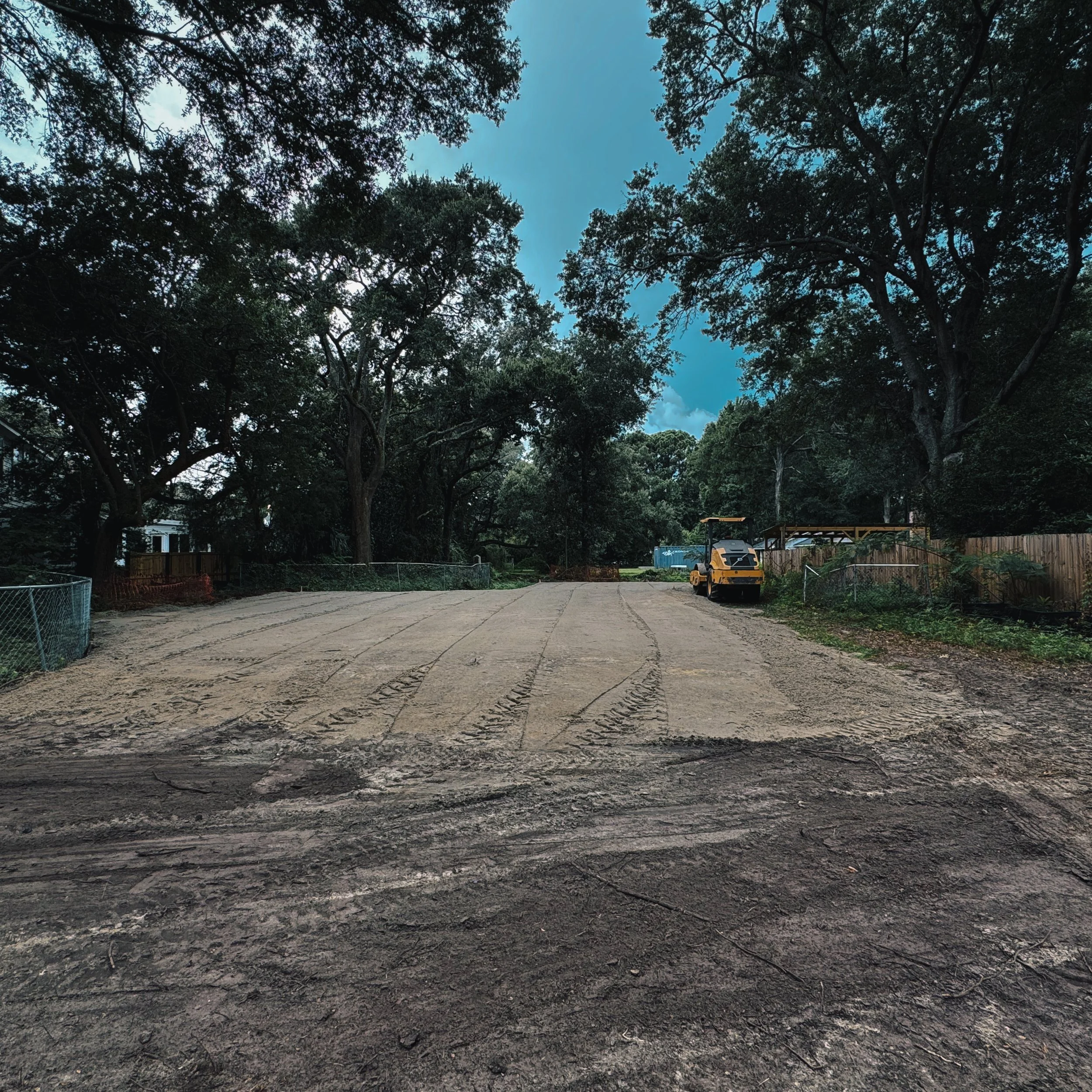 Large dirt area surrounded by trees with construction equipment on the right side, a yellow roller compactor and a fenced area, under a cloudy sky.