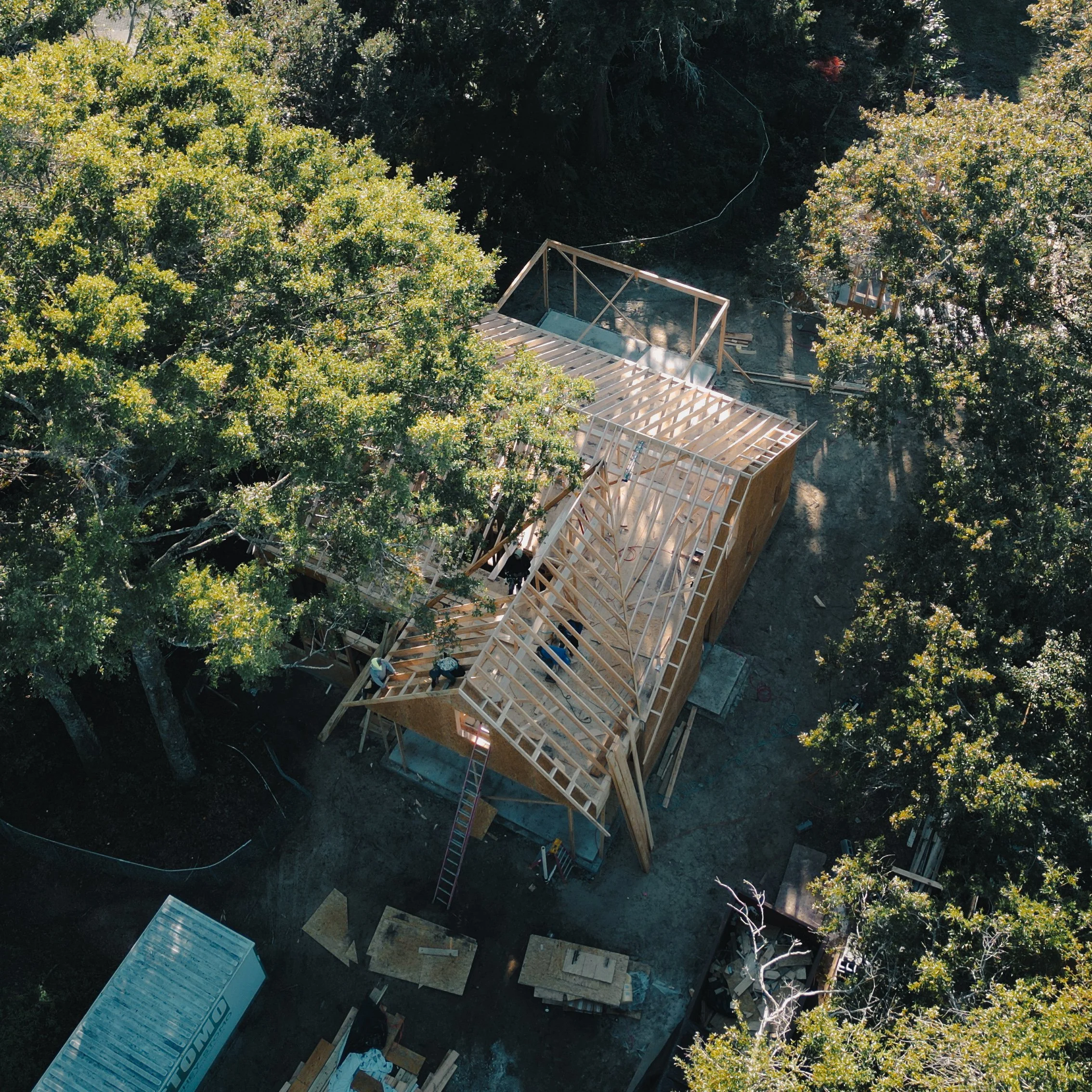 Aerial view of a house under construction surrounded by trees, showing the wooden framework and roof framing.