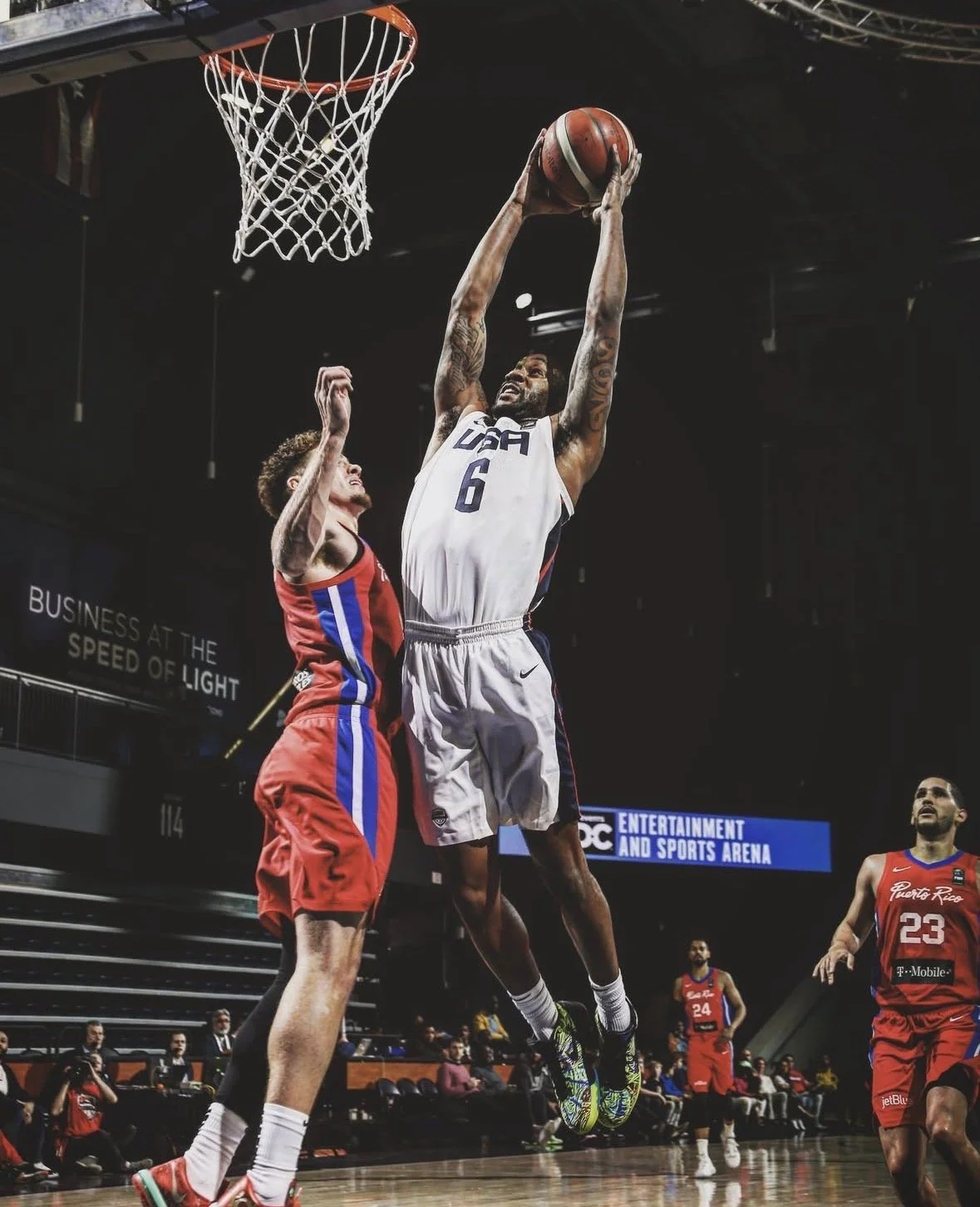 A basketball player in a USA jersey jumping to shoot the ball, while an opponent in a red and blue jersey tries to block him, during a game in an indoor arena.