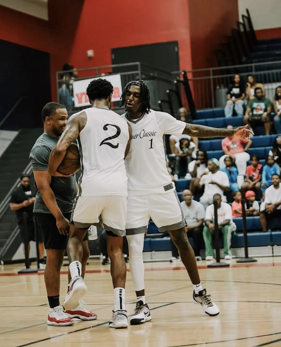 Two basketball players in white jerseys are talking on the court while standing near a coach or official. Spectators are seated in the background.