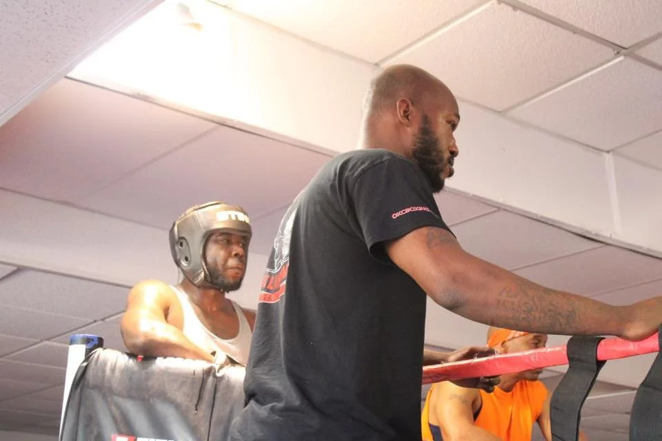 Two men in a gym lifting weights with a woman exercising in the background.