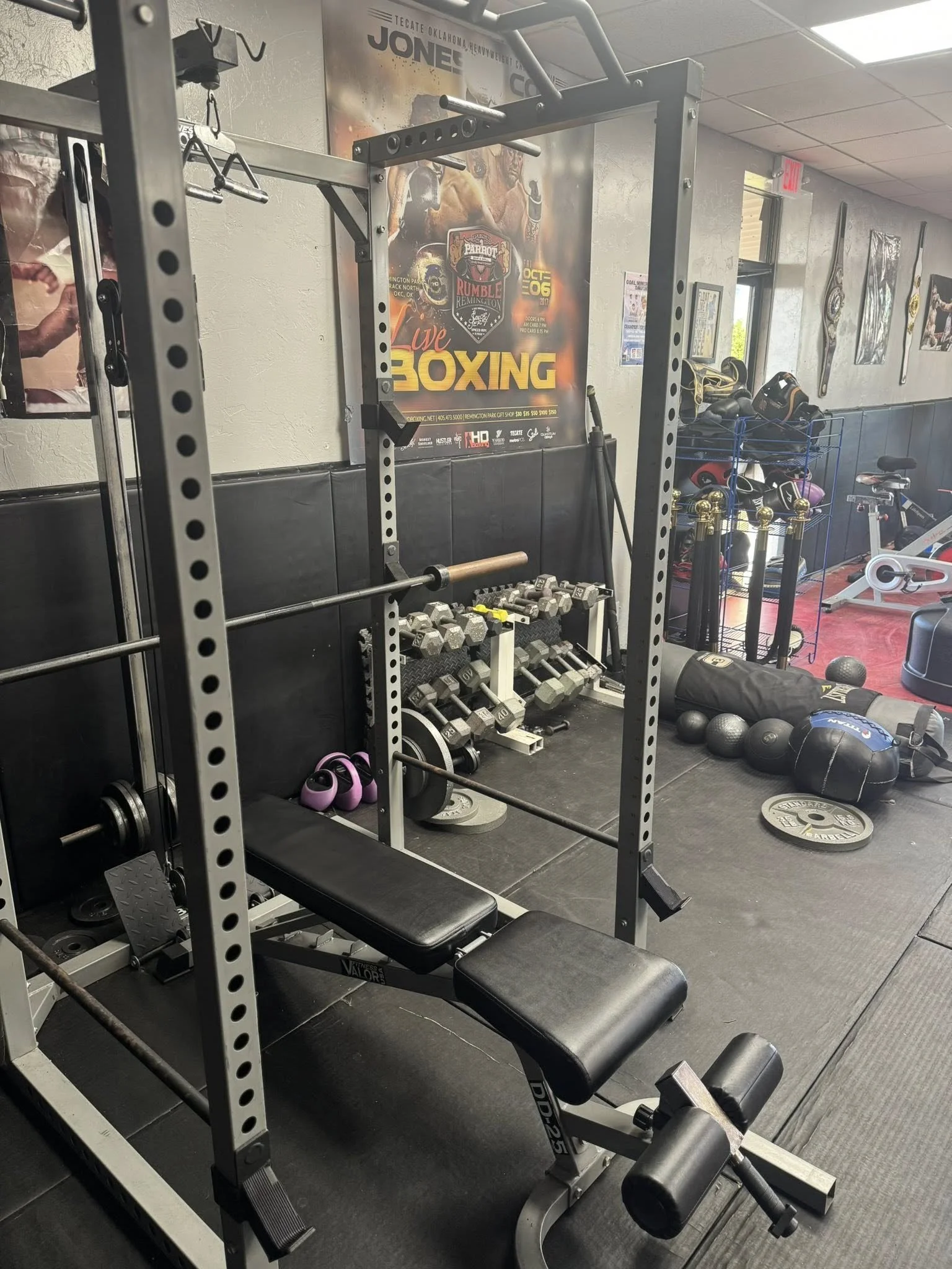 Gym workout area with weightlifting equipment, including a squat rack, dumbbells, kettlebells, medicine balls, and weight plates. There is a poster on the wall advertising a live boxing event.