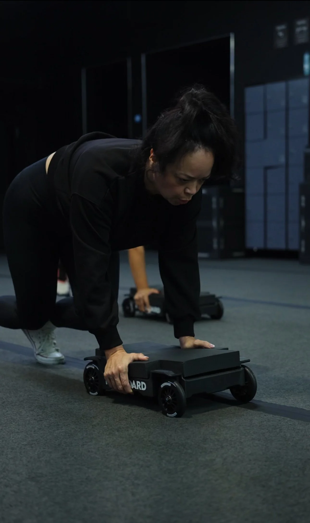 A woman in black athletic clothing is in a push-up position on a black skateboard with wheels, on a gym floor. She is focused, with her hands on the skateboard and her knees slightly bent, preparing for exercise.