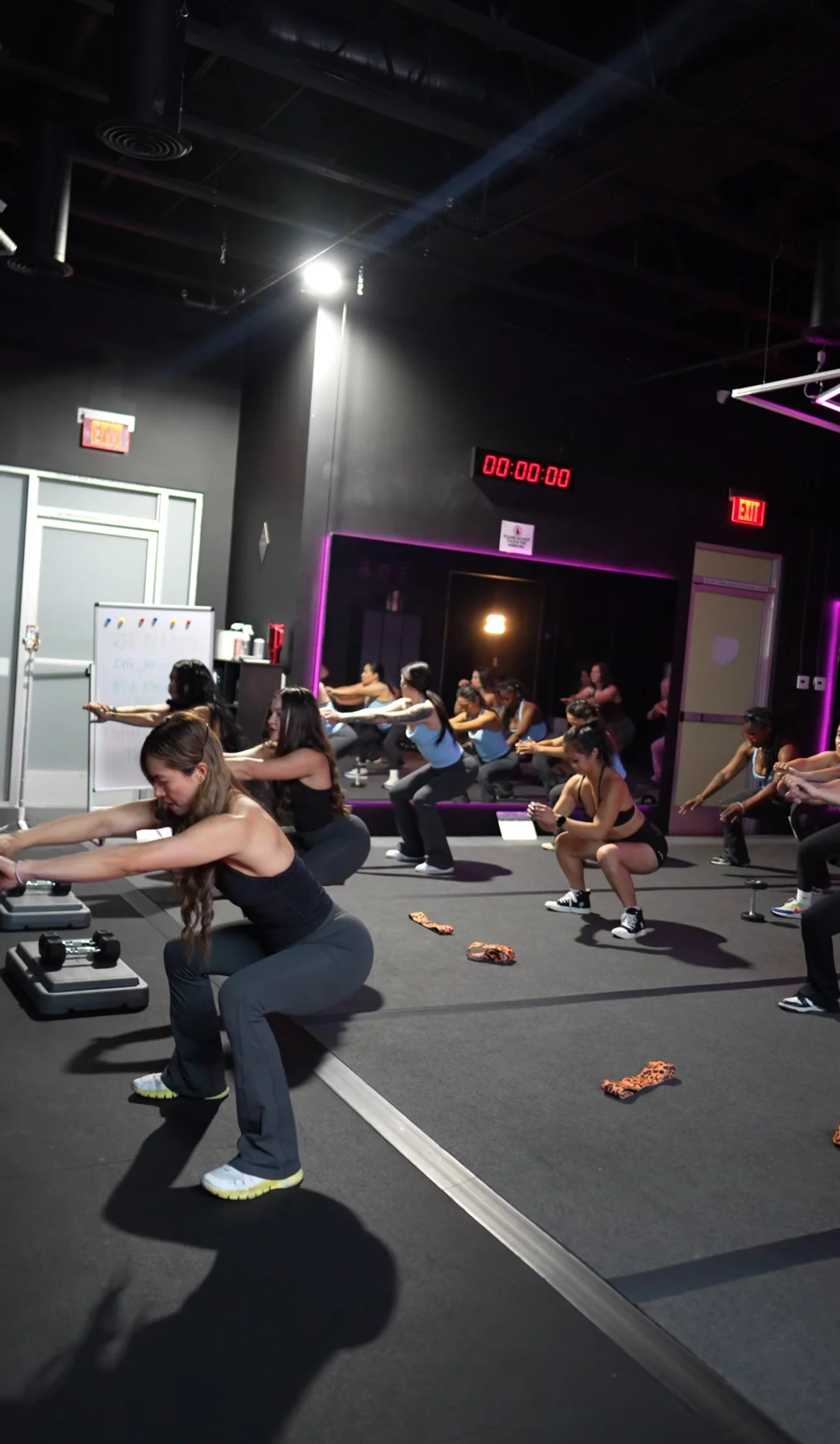 Women participating in a fitness class at a gym, doing squats with resistance bands, with a digital timer on the wall showing 00:00:00, and a mirror reflecting their actions in a dimly lit room with purple neon accents.