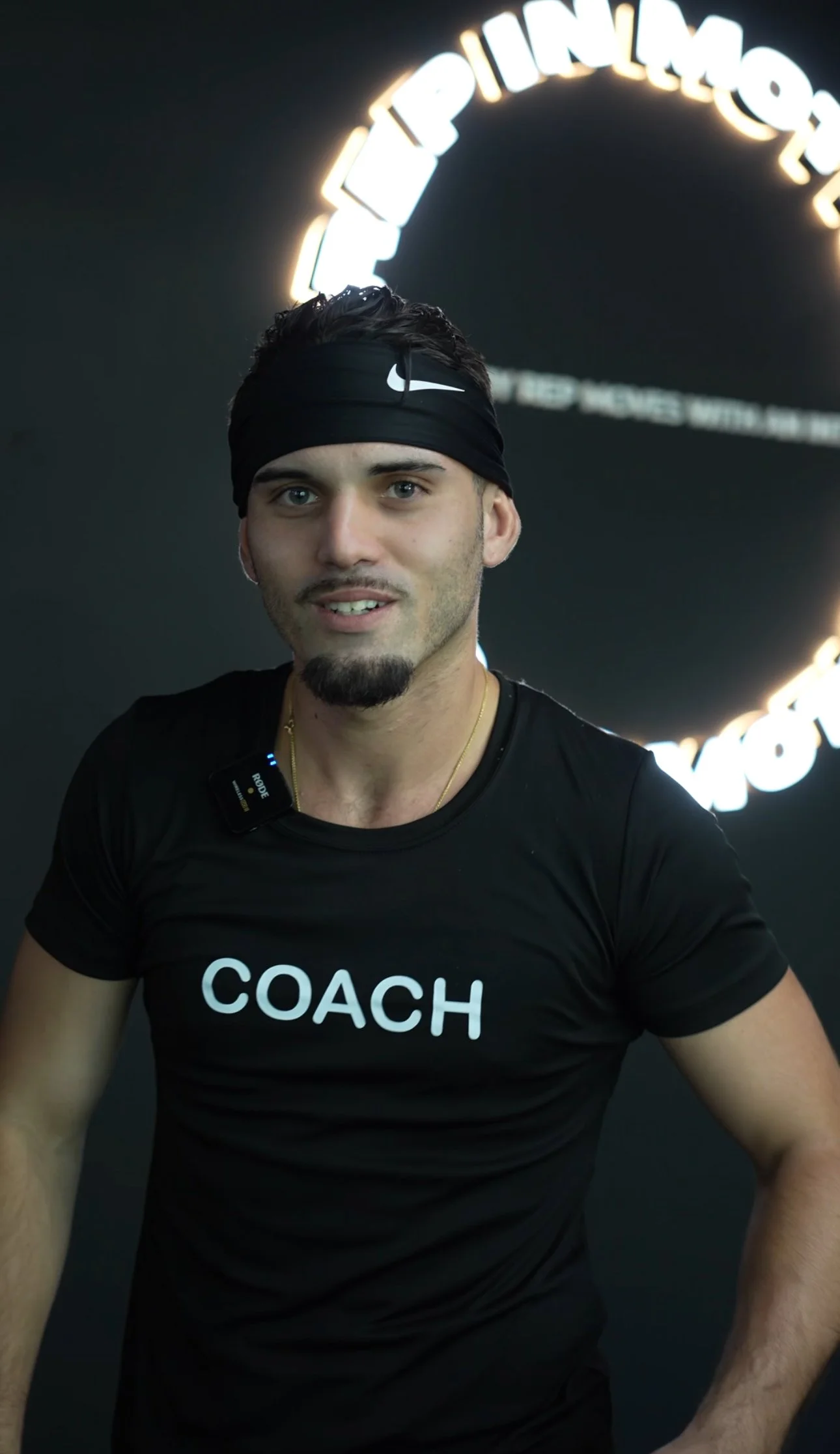 A young man wearing a black Nike headband and a black T-shirt with the word 'COACH' printed on it. He is standing in front of a dark background with glowing neon-style text that is partially visible.