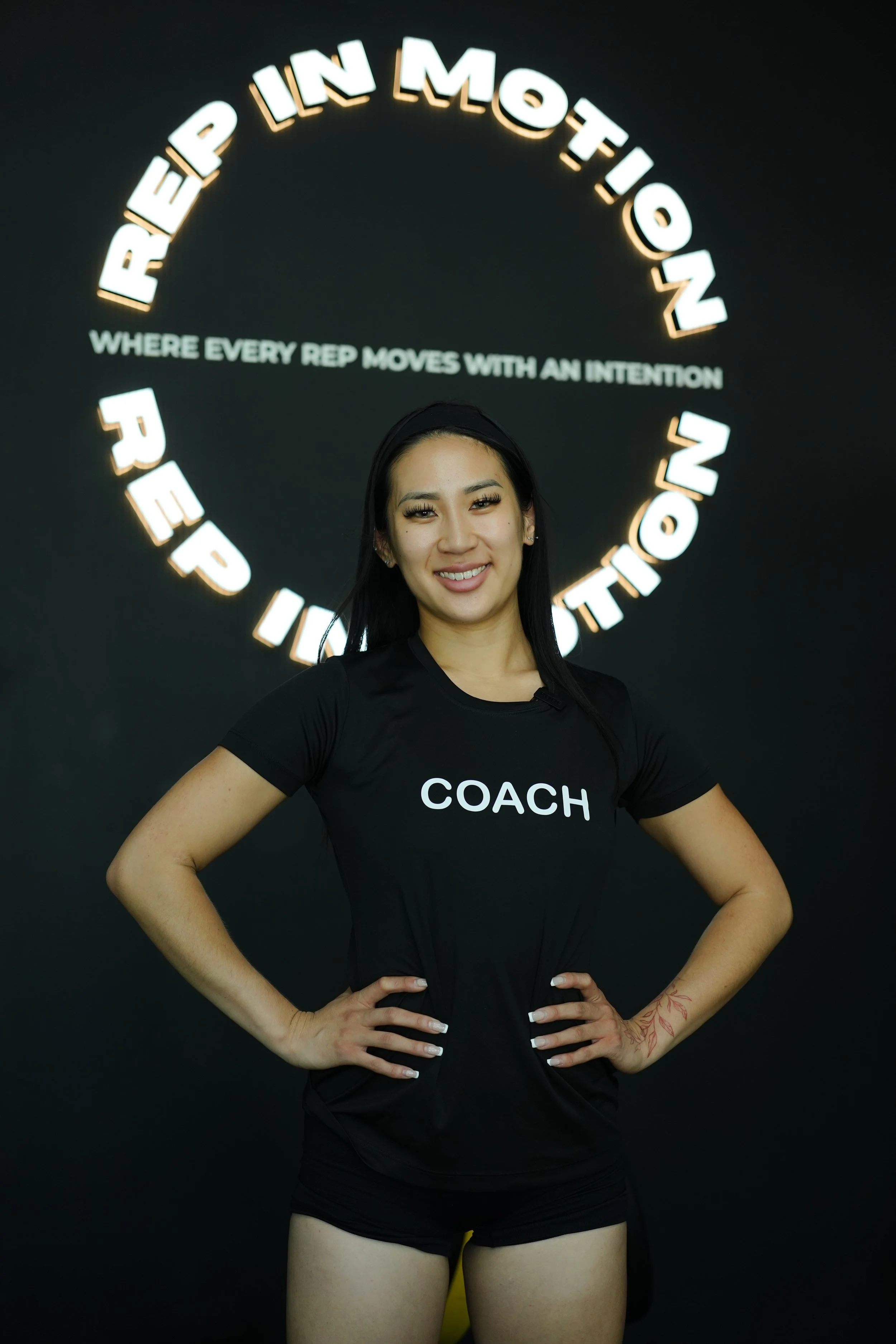 A woman standing in front of a black wall with illuminated text that reads "REP IN MOTION" in a circular arrangement and a smaller text "WHERE EVERY REP MOVES WITH AN INTENTION" underneath. She is wearing a black T-shirt with the word "COACH" printed on it and black shorts, smiling with hands on her hips.