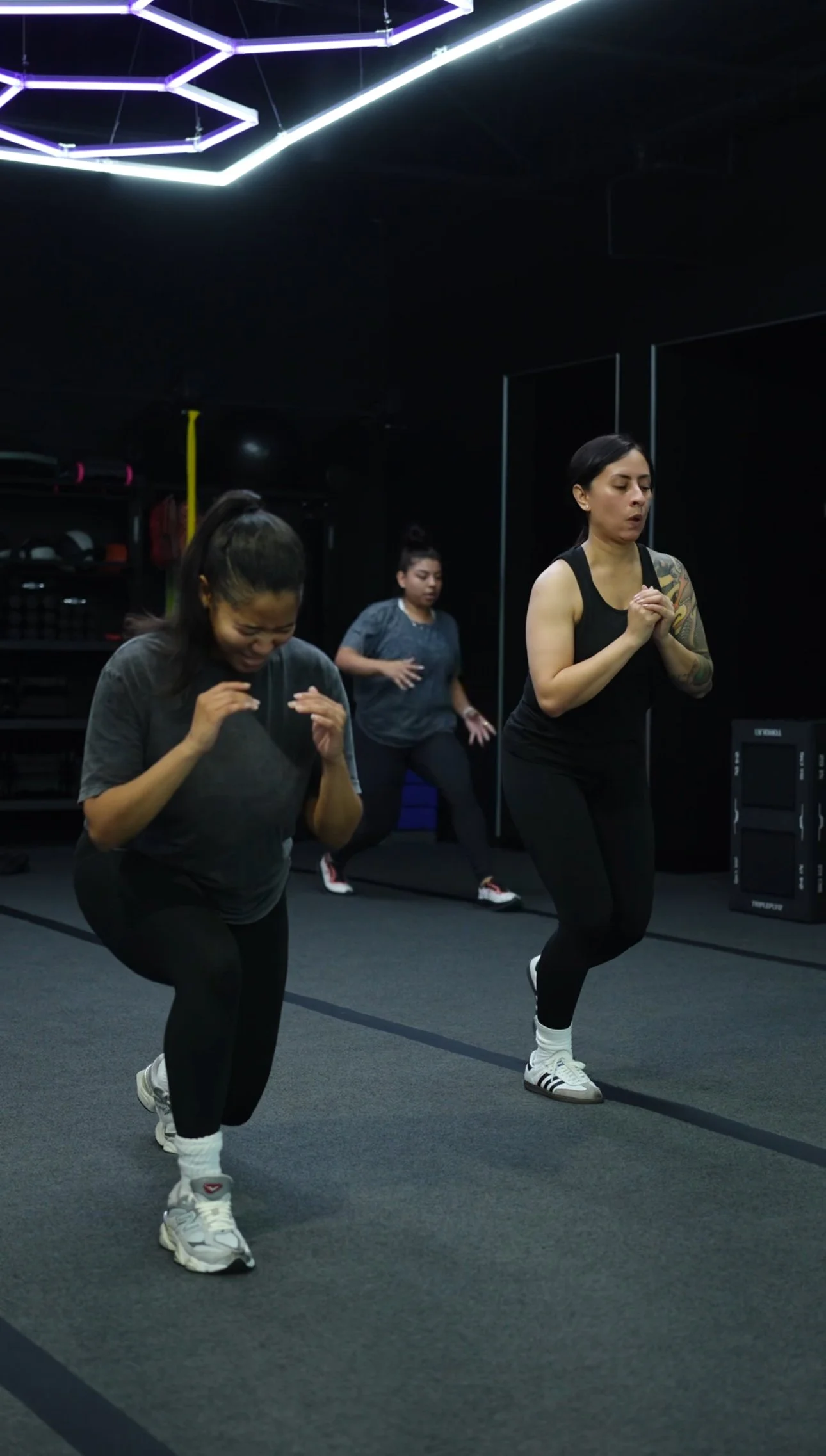 Three women exercising in a gym, performing lunges while wearing athletic clothing and sneakers.