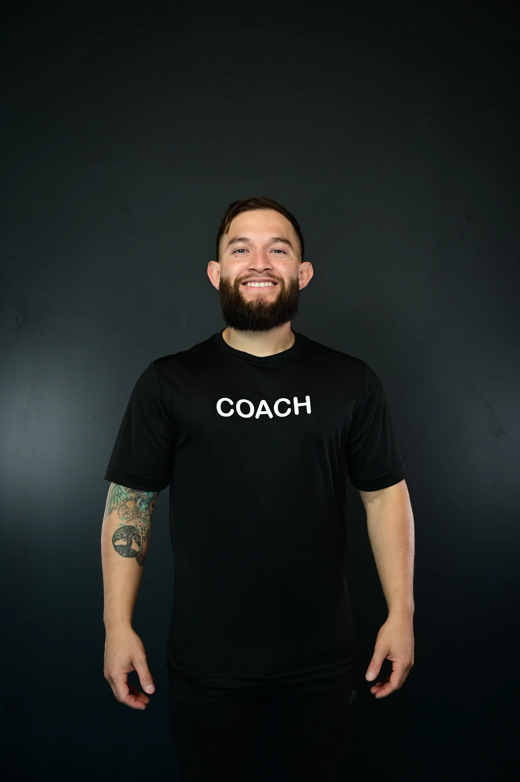 A smiling man with a beard wearing a black T-shirt with 'COACH' written on it, standing against a dark background.