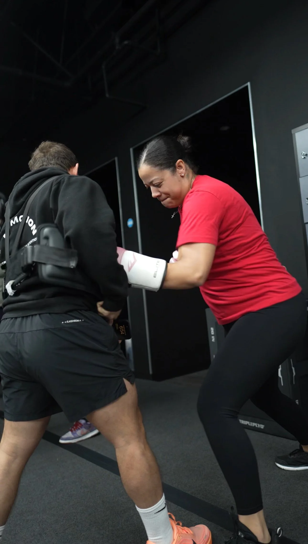 A woman in a red shirt and black leggings practicing boxing with a man in dark athletic clothing wearing boxing gloves in a gym.