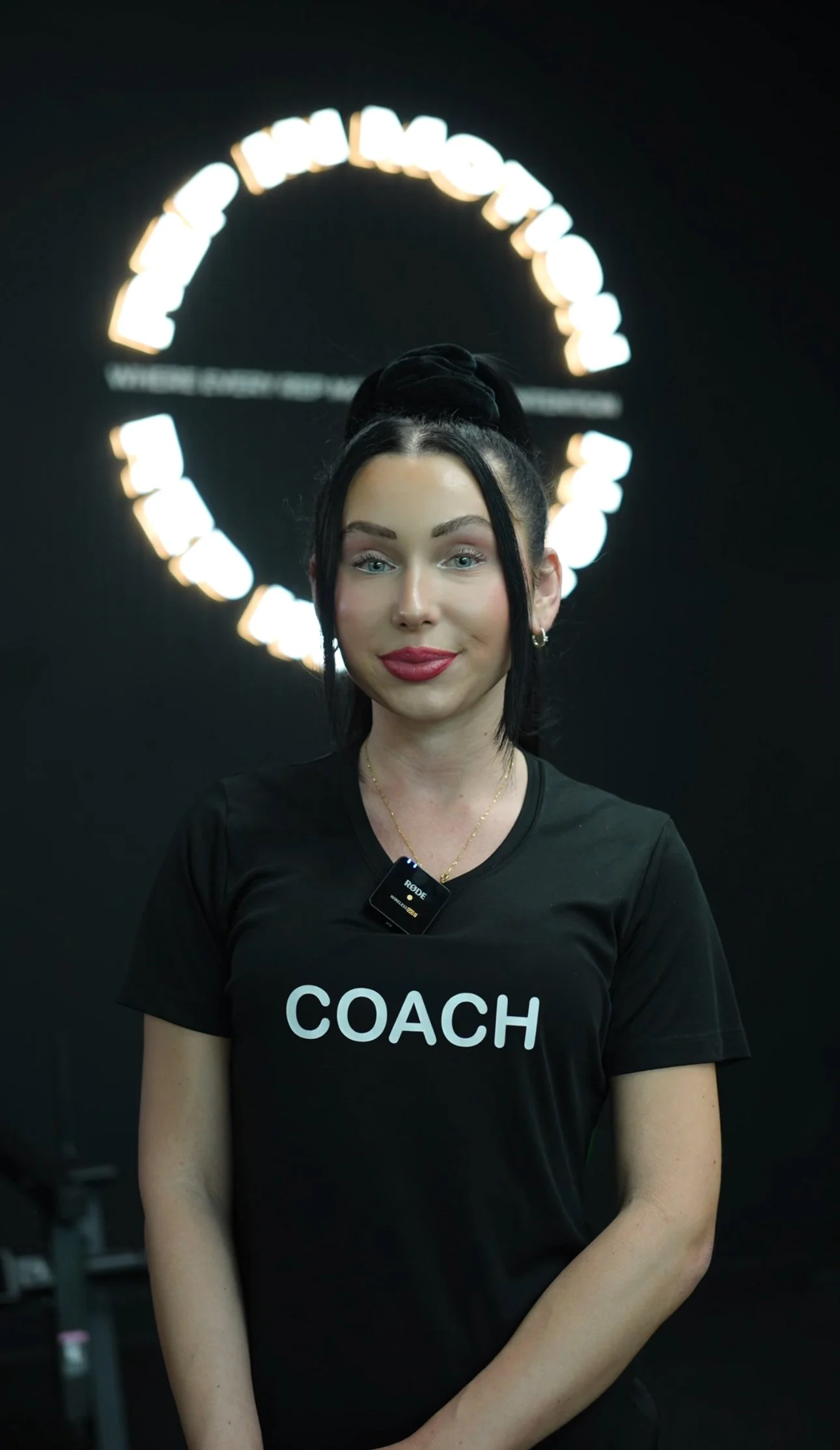 A woman standing in front of a black background with a glowing circular logo, wearing a black t-shirt with 'COACH' printed on it, and a microphone clipped to her shirt.