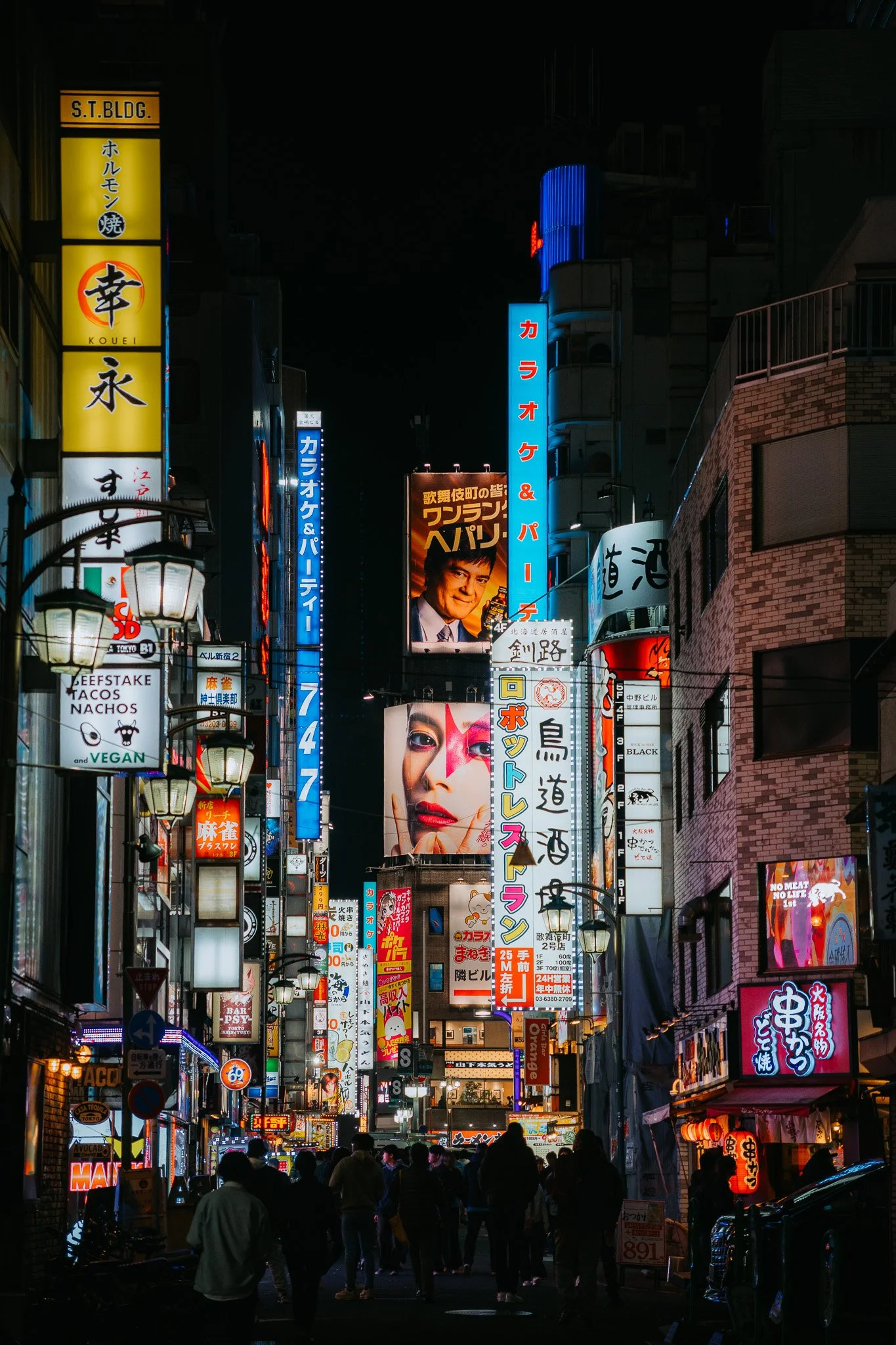 Nighttime street scene in Shinjuku with bright neon signs, billboards, and people walking on the street.