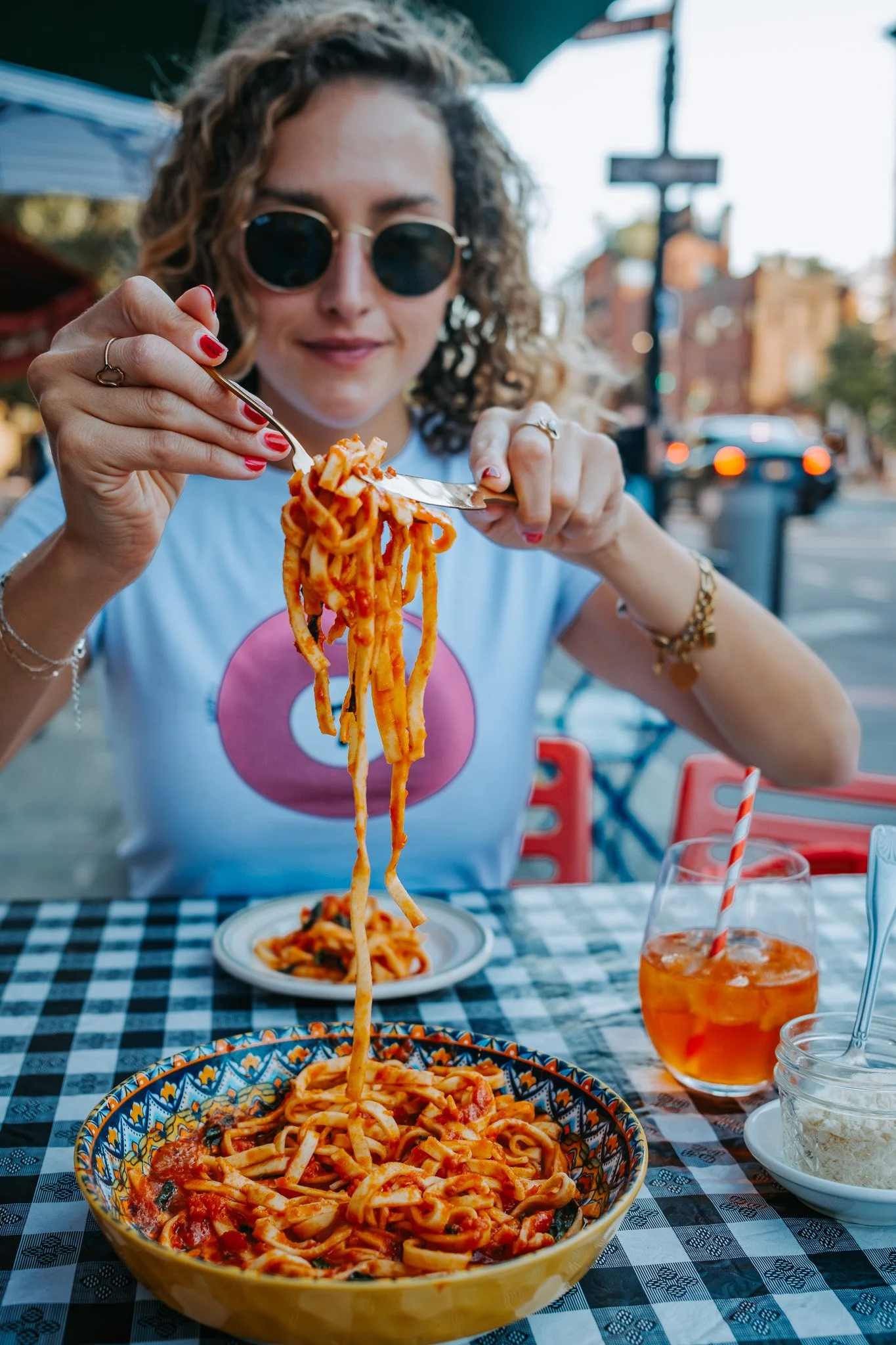 A woman with curly hair, sunglasses, and red nail polish sitting at an outdoor table, enjoying a plate of spaghetti with tomato sauce and a drink with an orange straw.