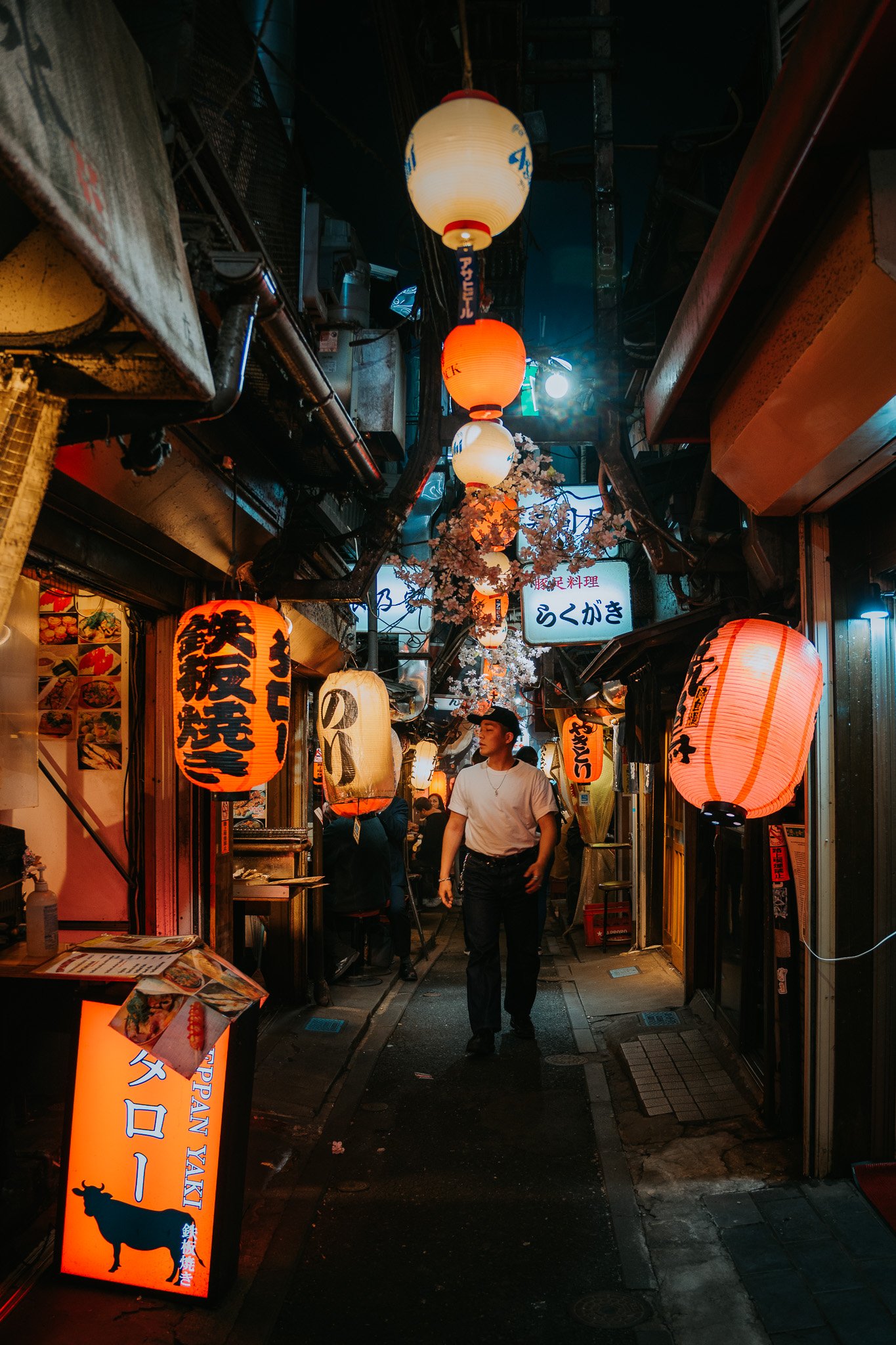 Night scene of the famous narrow Japanese alley, Omiode Yokocho (Memory Lane) decorated with illuminated paper lanterns, hanging cherry blossom branches, and signs for restaurants, with a man walking through.