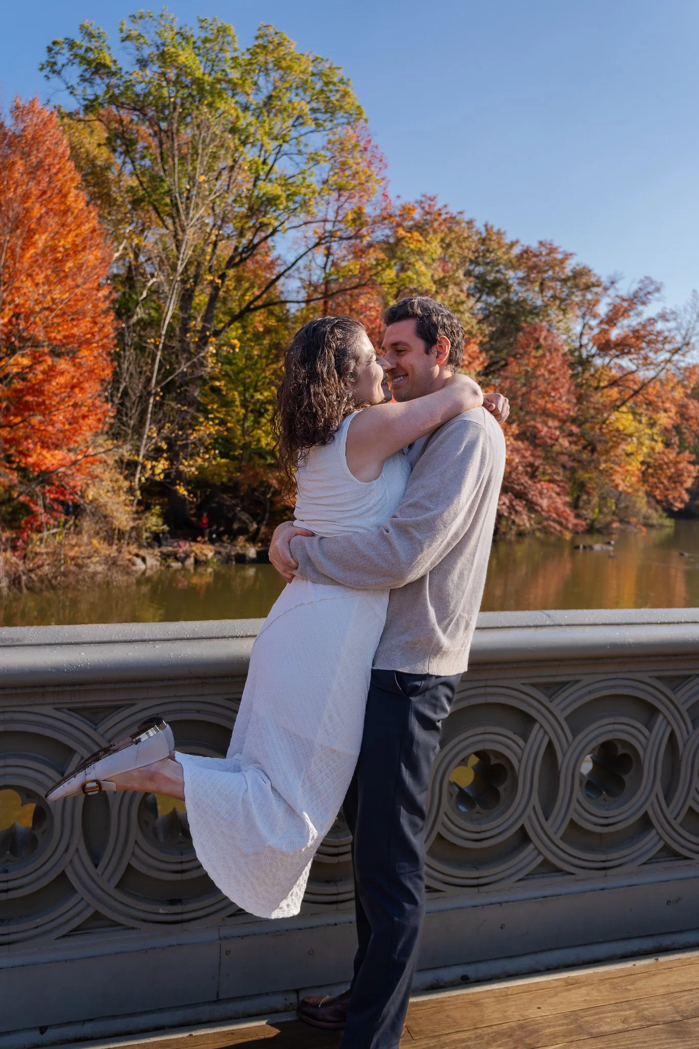 A couple embracing on a bridge during autumn, with colorful fall trees and a river in the background.