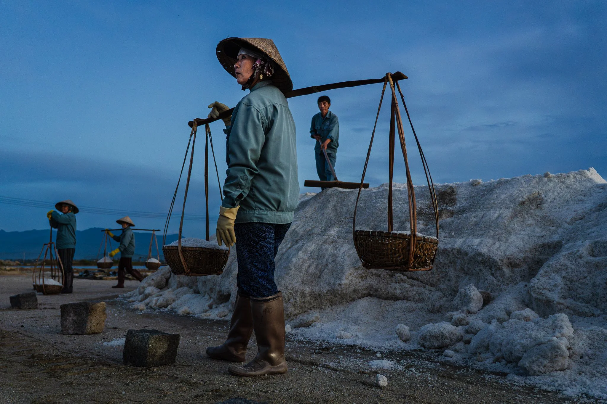 Four Vietnamese workers carrying baskets on their shoulders while walking along a salt field with large salt piles, wearing hats, gloves, and boots during dusk.