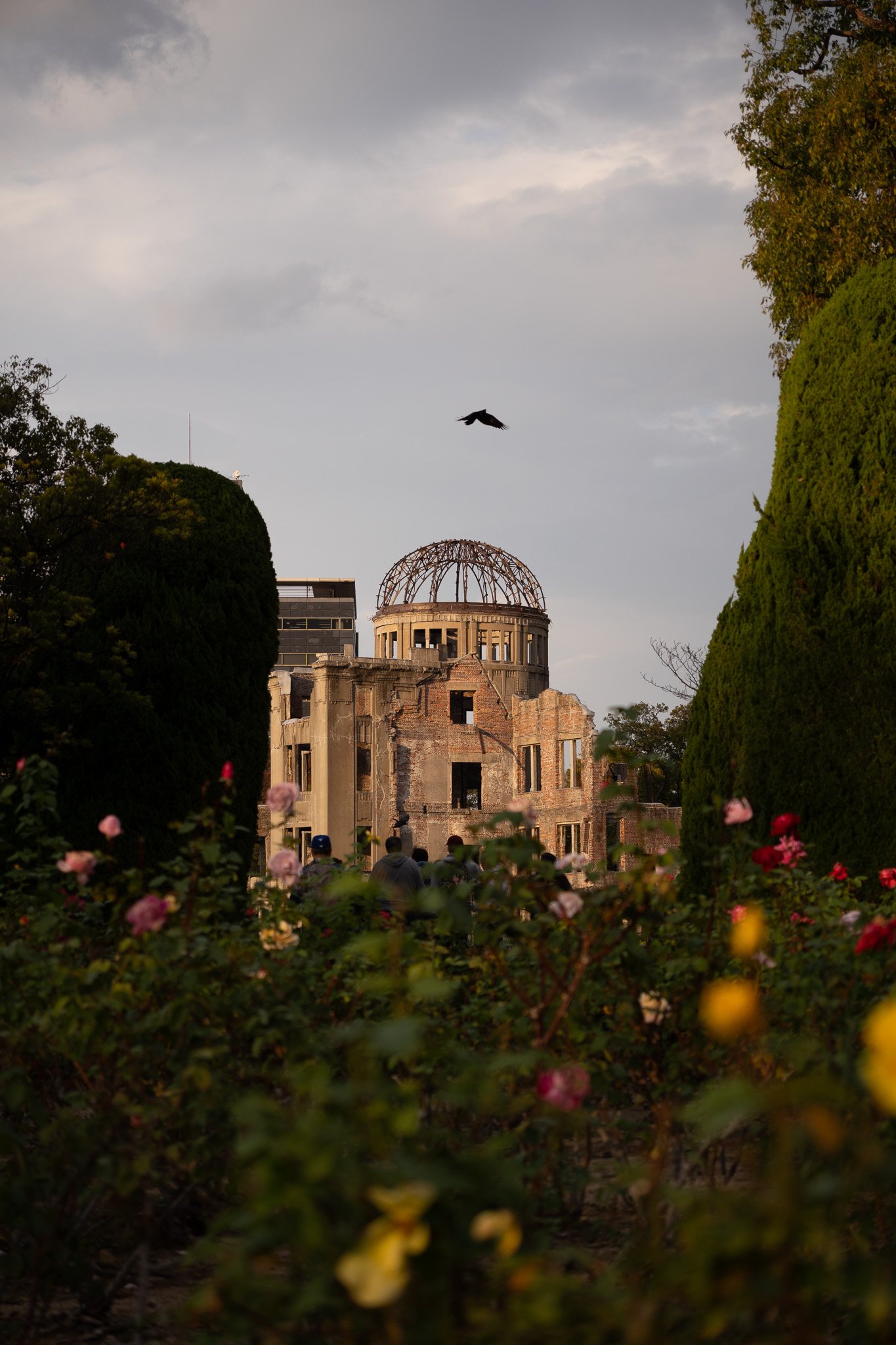 The Hiroshima Peace Memorial, also known as the Atomic Bomb Dome, with colorful flowers in foreground and a bird flying above, under a cloudy sky.