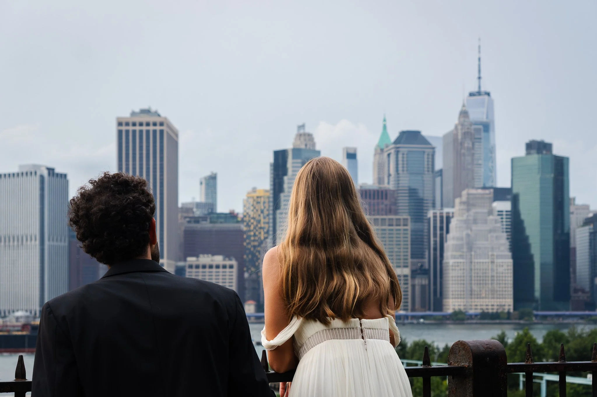 A bride and groom look at the Manhattan city skyline with tall buildings across a river from Brooklyn heights.