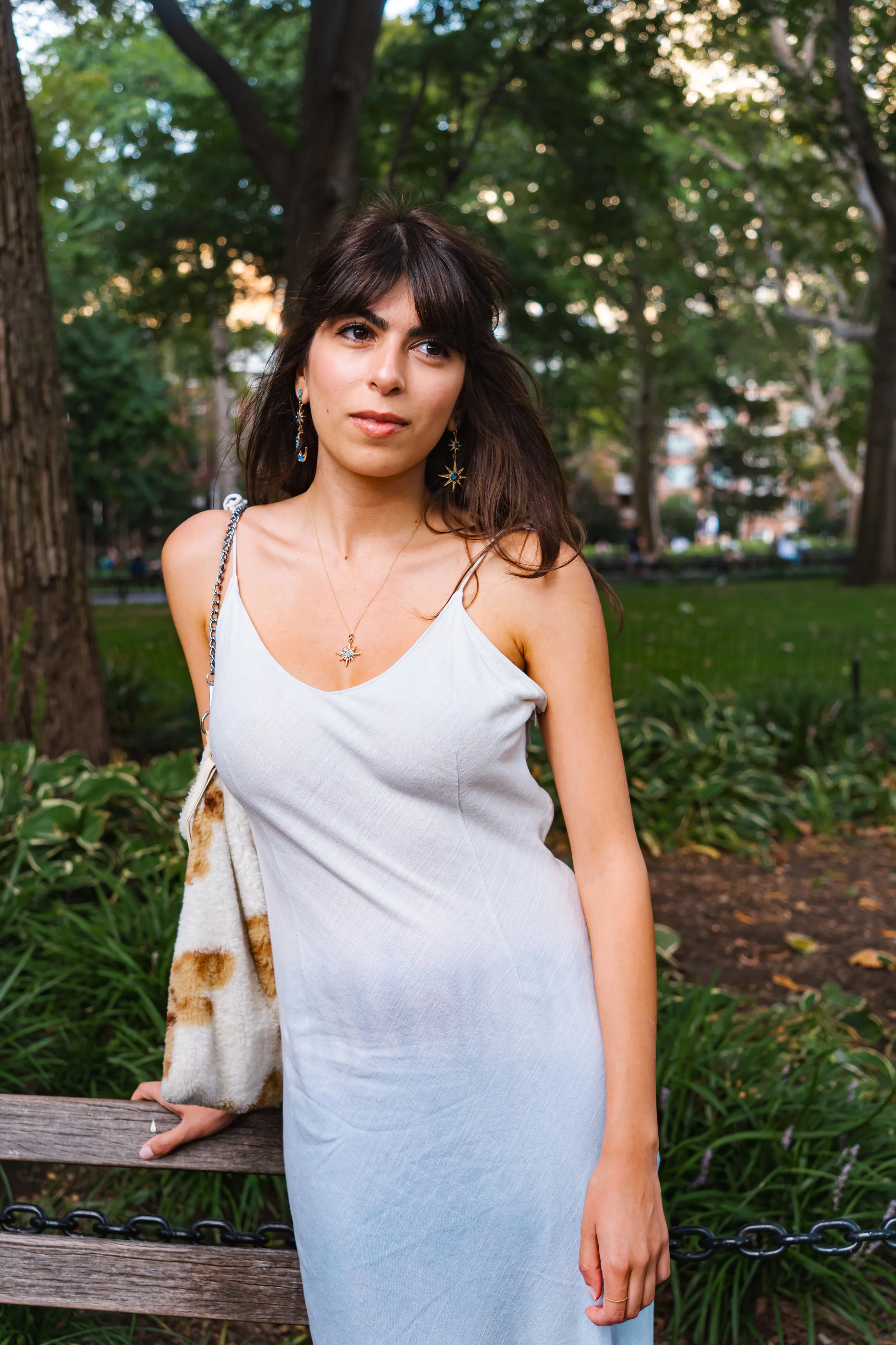 A young woman with dark hair and brown eyes stands outdoors in Washington Square Park, wearing a white sleeveless dress and jewelry, with her left hand resting on a wooden bench.