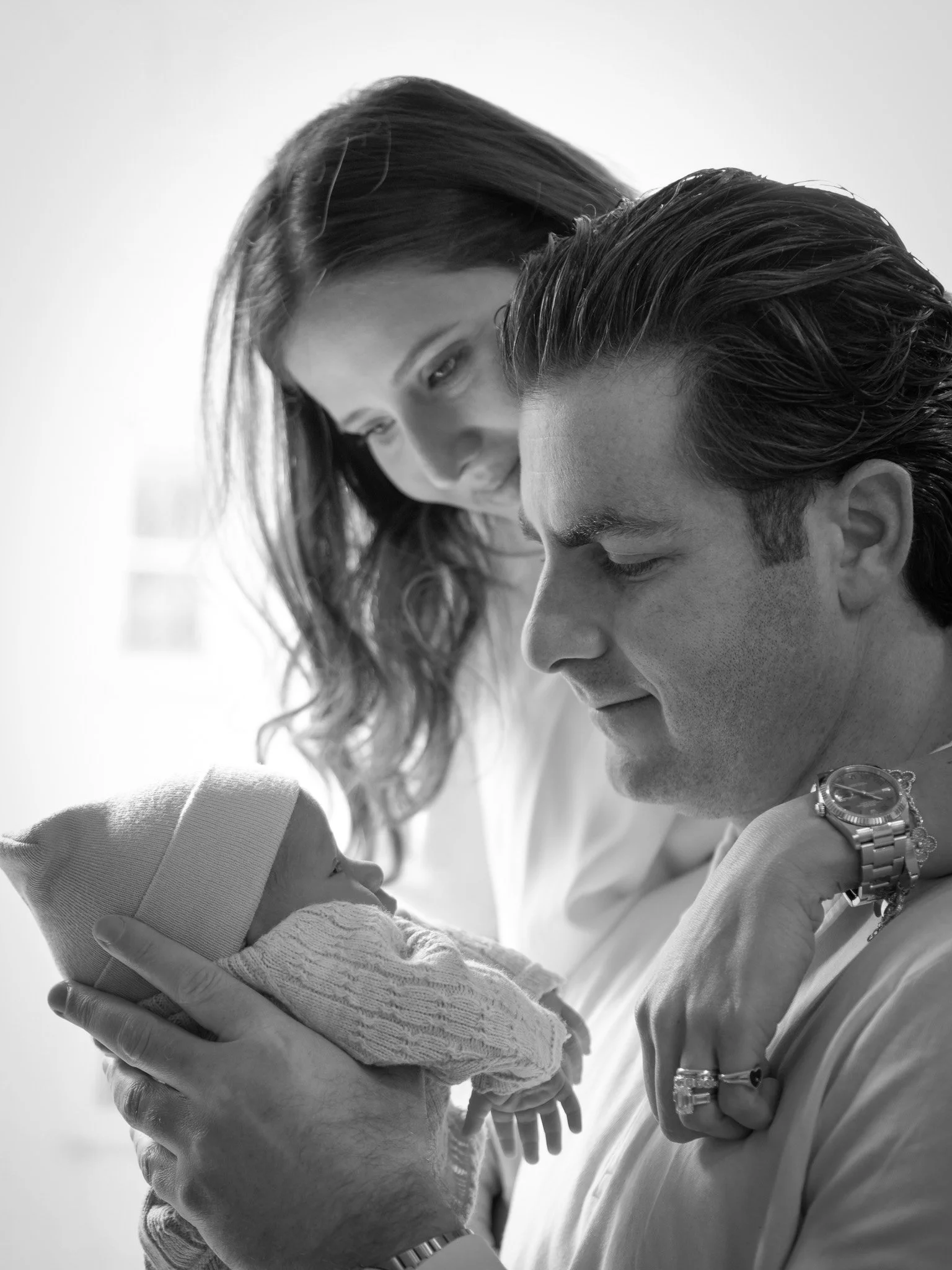 A woman and a man look lovingly at a newborn baby, with the woman leaning over the man who is holding the baby in his hands.