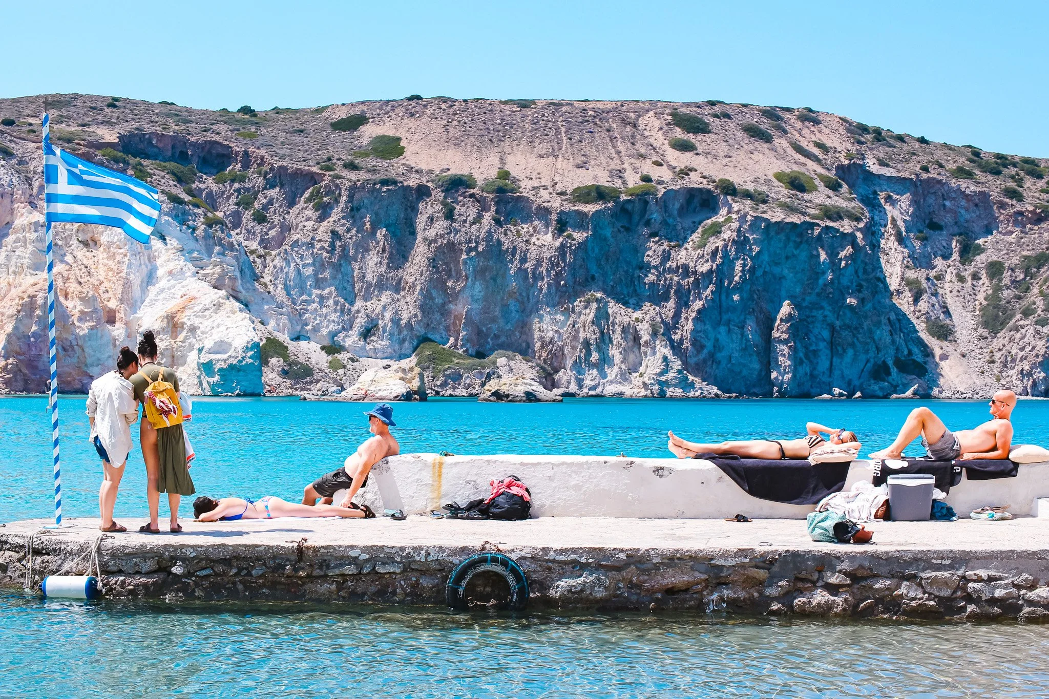 People relaxing and enjoying the sunny weather on a concrete pier by the water with rocky cliffs and a Greek flag in the background