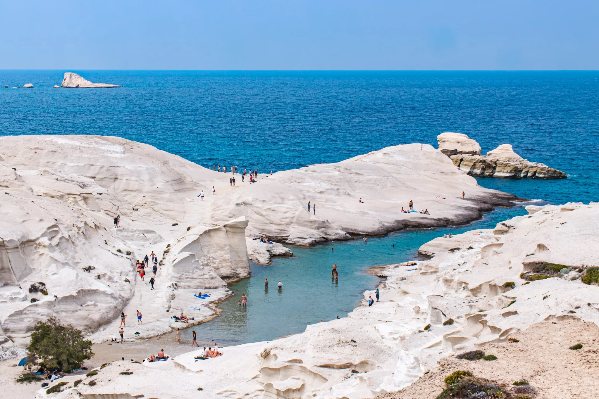 Sarakiniko Beach with white rocky formations, people swimming and sunbathing, blue sea, clear sky.