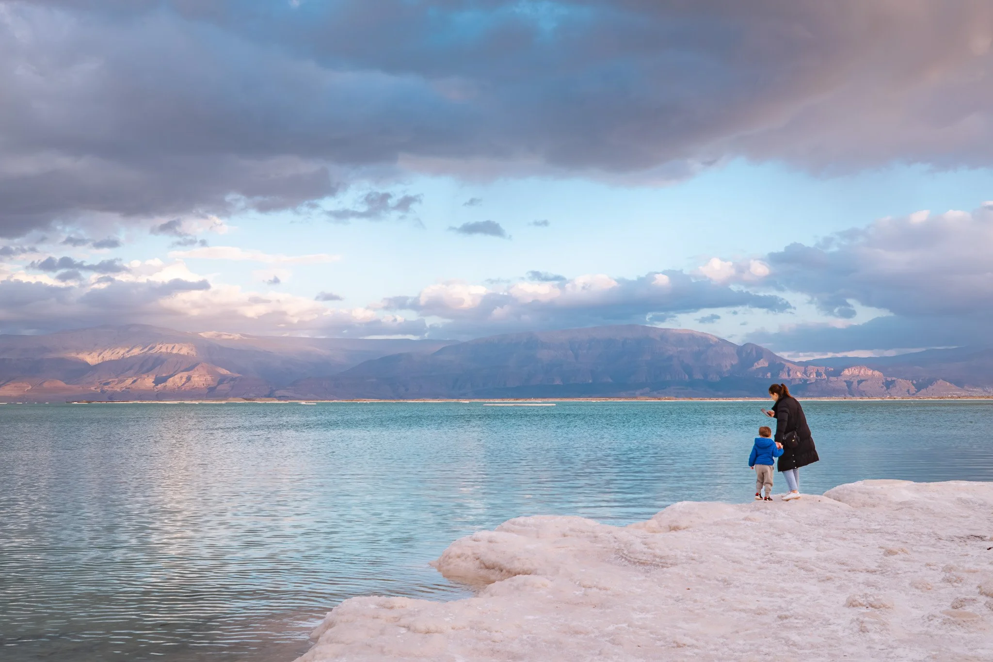 A woman and a young child standing on a salt flat at the Dead Sea, with mountains and a cloudy sky in the background.